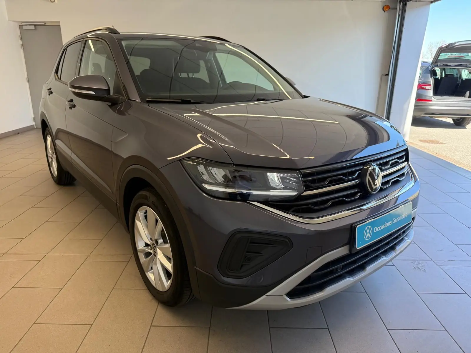 Volkswagen T-Cross 2025 gris cendre en 3/4 avant droit, avec calandre noire et détails chromés visibles en showroom.