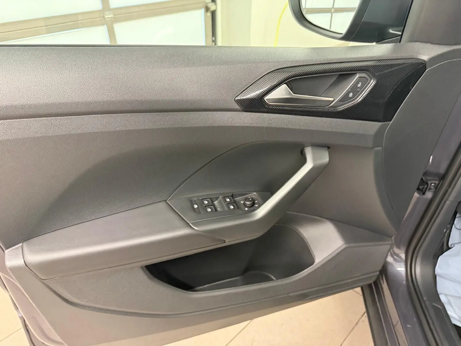 Vue intérieure de la porte avant gauche du Volkswagen T-Cross gris cendre, avec commandes des vitres et poignée intégrée.