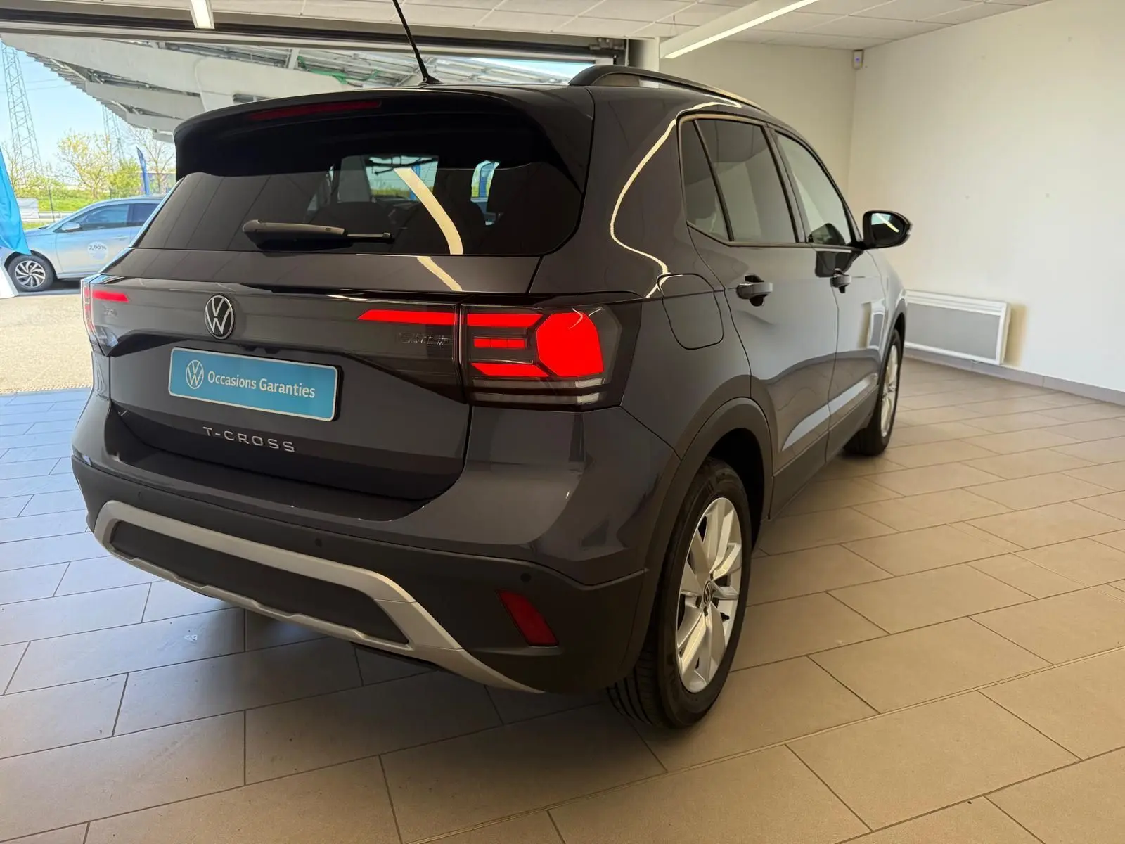 Volkswagen T-Cross gris cendre vue 3/4 arrière droit en intérieur, feux arrière allumés et logo VW visible.
