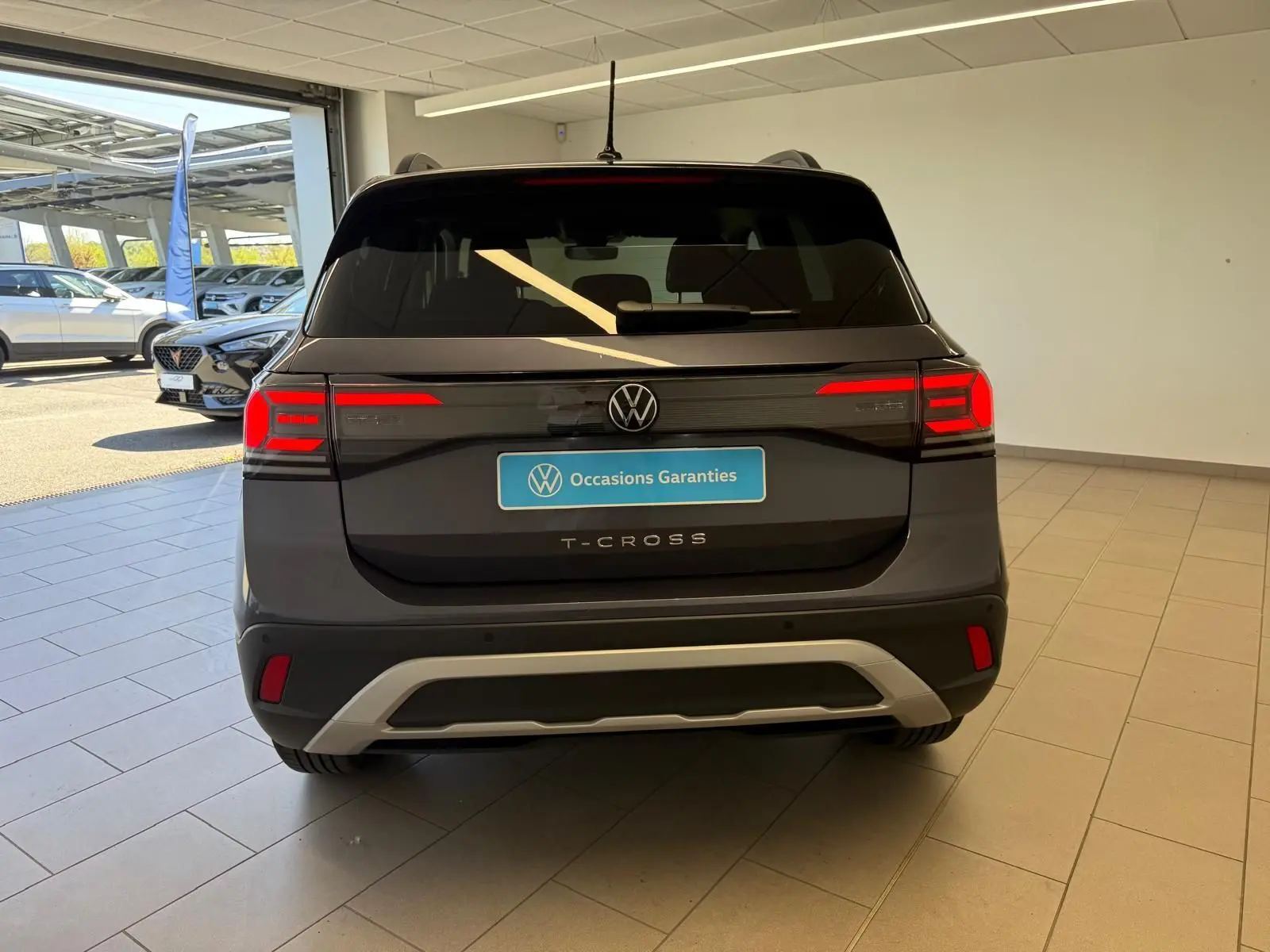 Vue arrière d'un Volkswagen T-Cross gris cendre 2025 avec feux arrière allumés et logo VW visible.