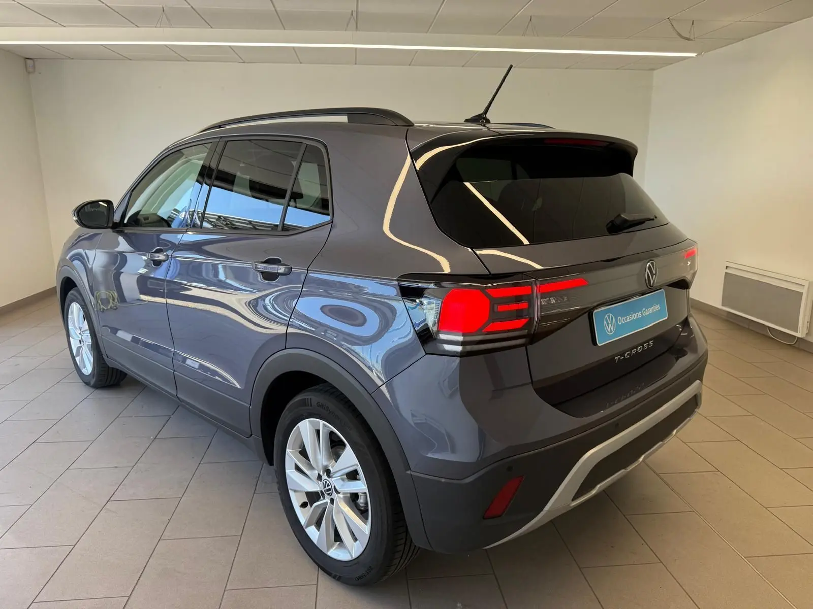 Volkswagen T-Cross gris cendre vu en 3/4 arrière droit, avec feux arrière allumés et jantes argentées.