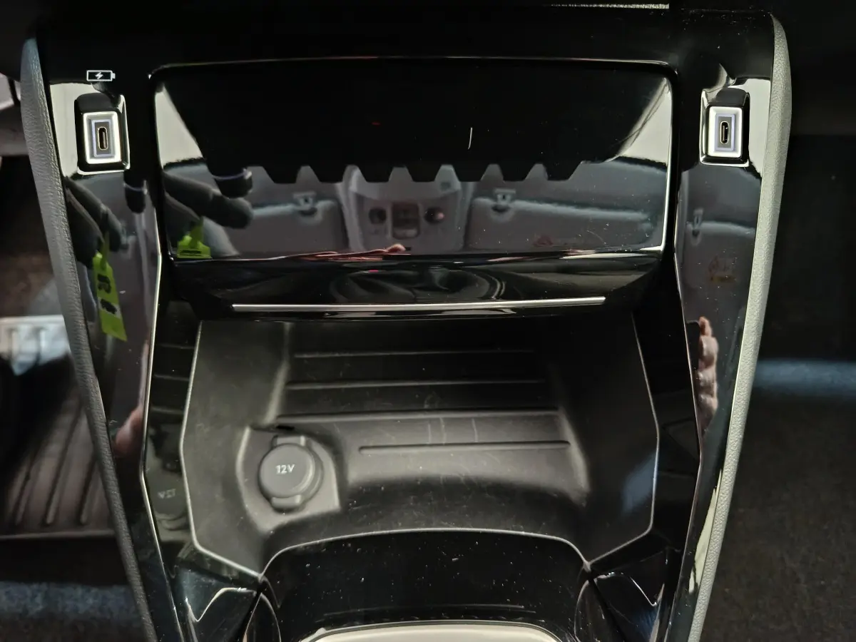 Gros plan sur la console centrale noire brillante de la Peugeot 208 PureTech 100 S&S Allure avec prise 12V et ports USB.