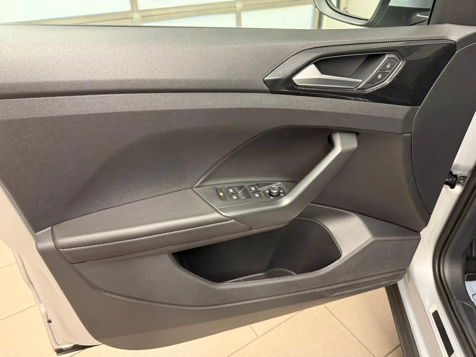 Vue intérieure de la porte avant gauche du Volkswagen T-Cross 2024 en Reflet d'Argent, avec commandes électriques et poignée intégrée.