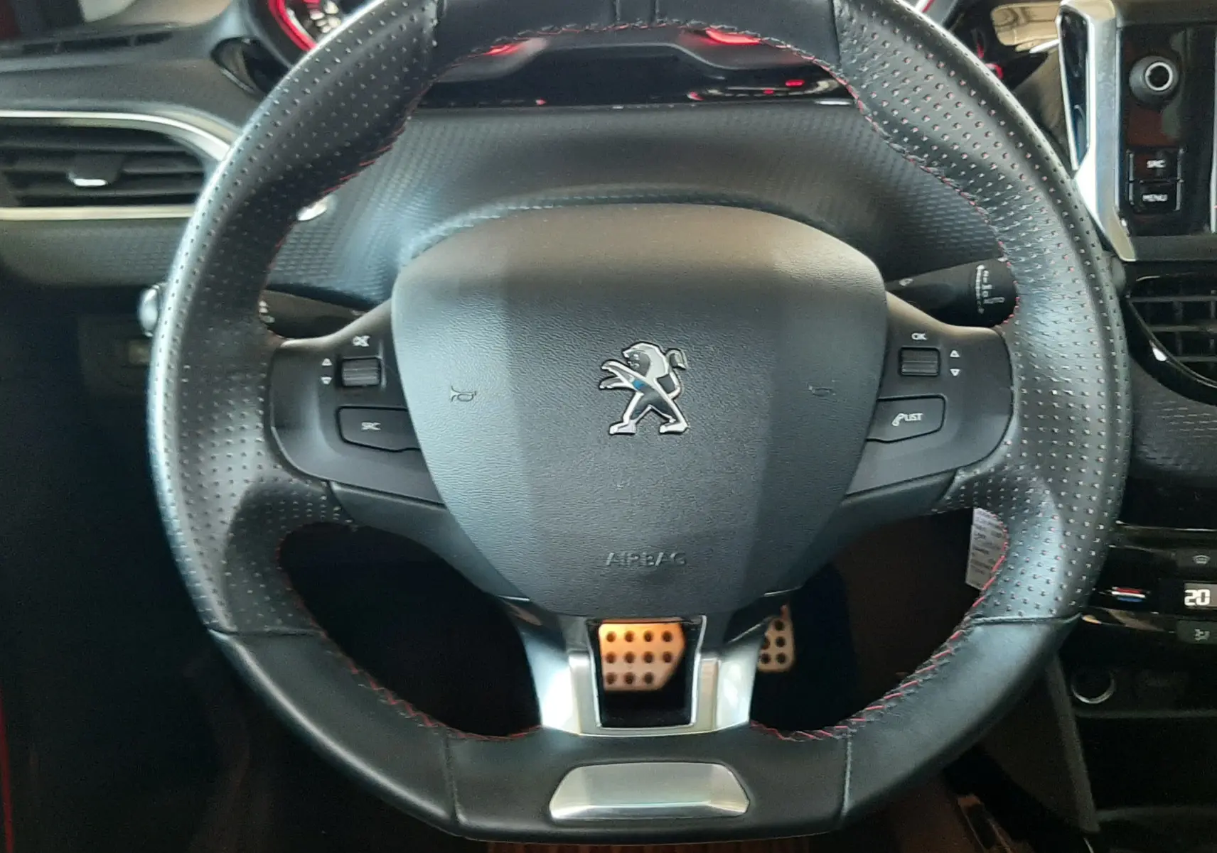 Volant en cuir perforé avec surpiqûres rouges et logo Peugeot, tableau de bord et pédales sportives visibles en arrière-plan.