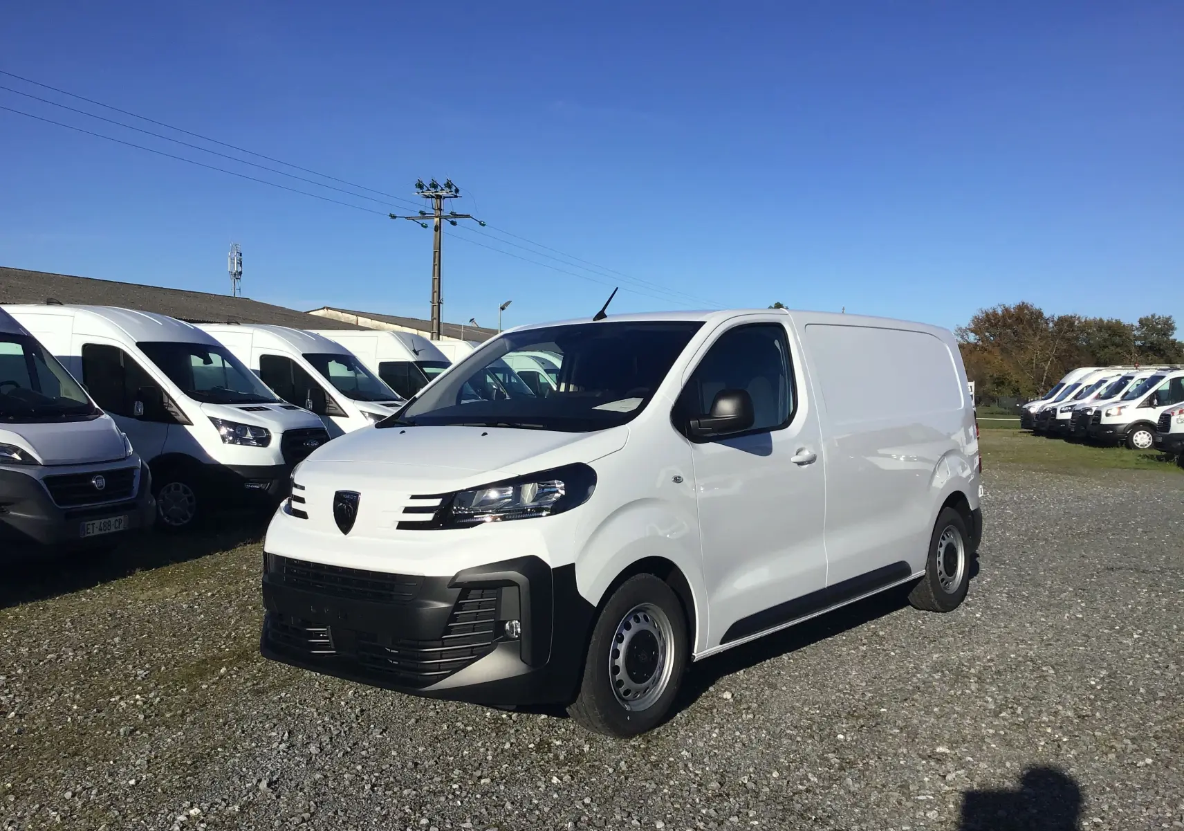 Fourgon Peugeot Expert blanc en 3/4 avant droit, stationné sur un terrain avec plusieurs véhicules utilitaires en arrière-plan.