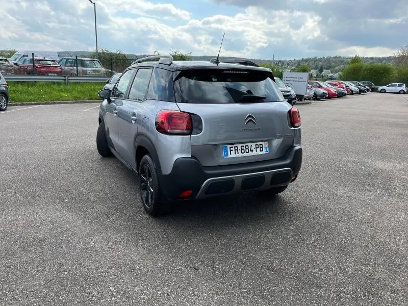 Vue 3/4 arrière droite d'un Citroën C3 Aircross gris acier avec toit noir et barres de toit sur parking.