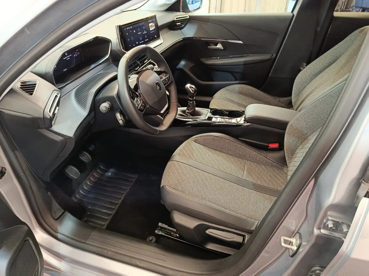 Intérieur avant droit de la Peugeot 208 gris clair 2025, volant compact, écran tactile central et sièges tissu gris.