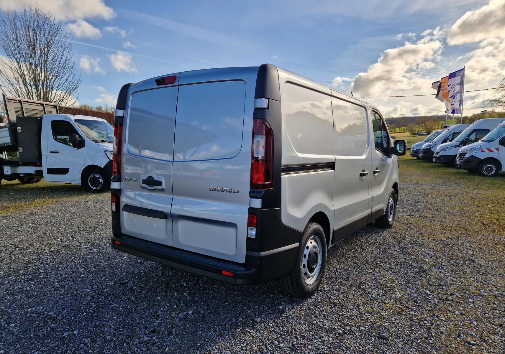 Vue 3/4 arrière droite d’un Renault Trafic Fourgon gris Highland garé sur un sol gravillonné avec plusieurs autres utilitaires en arrière-plan.