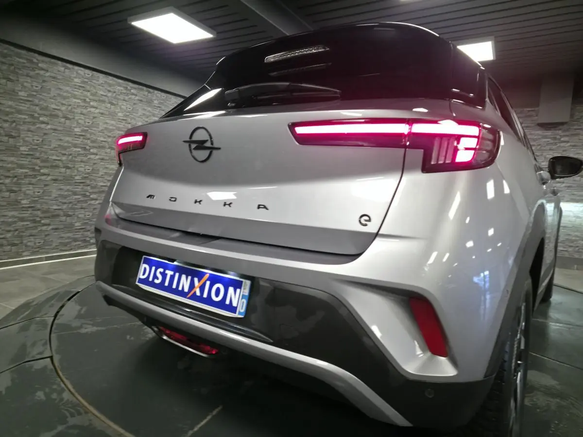 Vue 3/4 arrière droite d'un Opel Mokka-e gris avec feux arrière LED allumés et logo noir sur coffre.