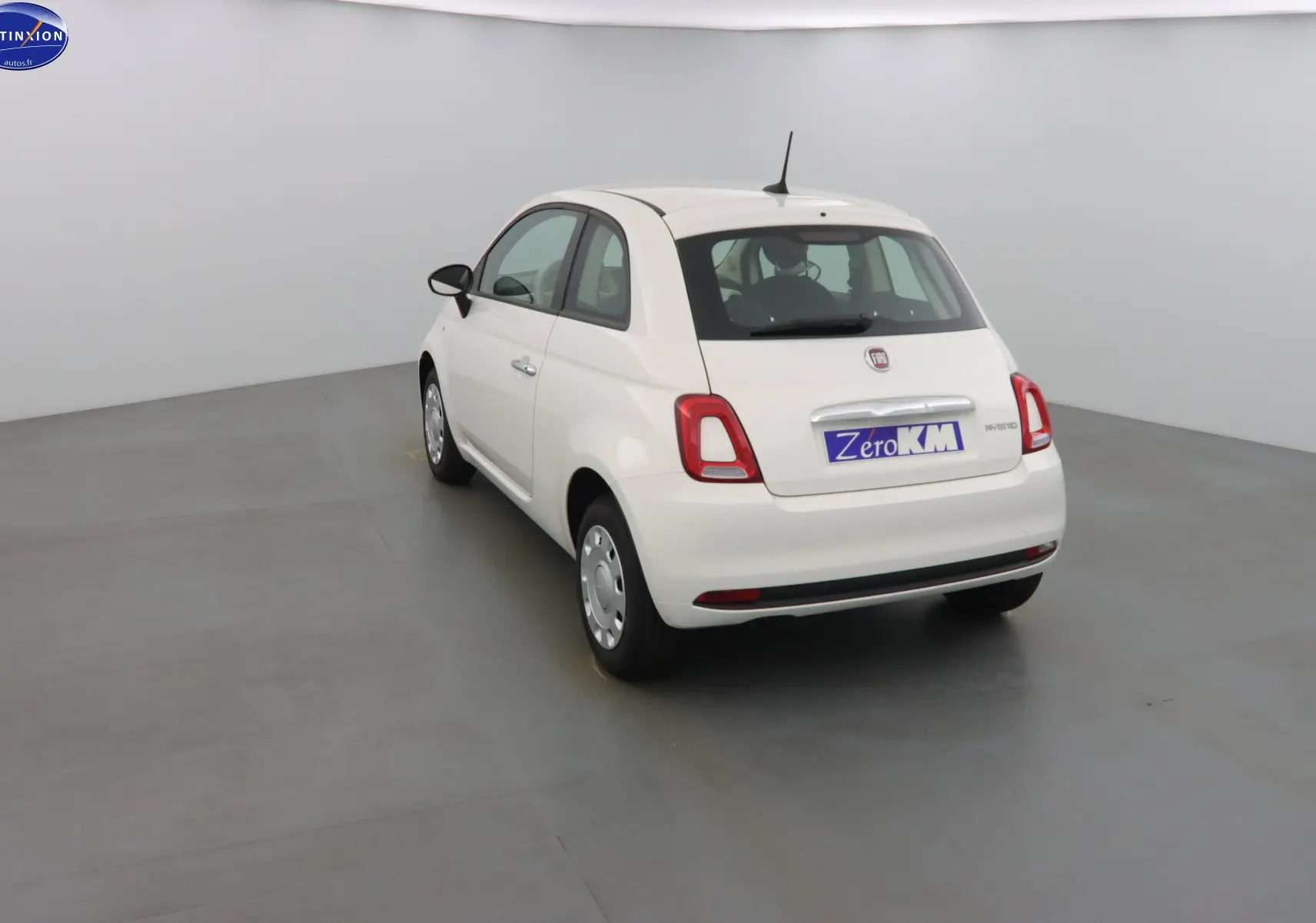 Fiat 500 blanc gelato vue 3/4 arrière droit dans un studio, avec badge hybride et plaque Zero KM.