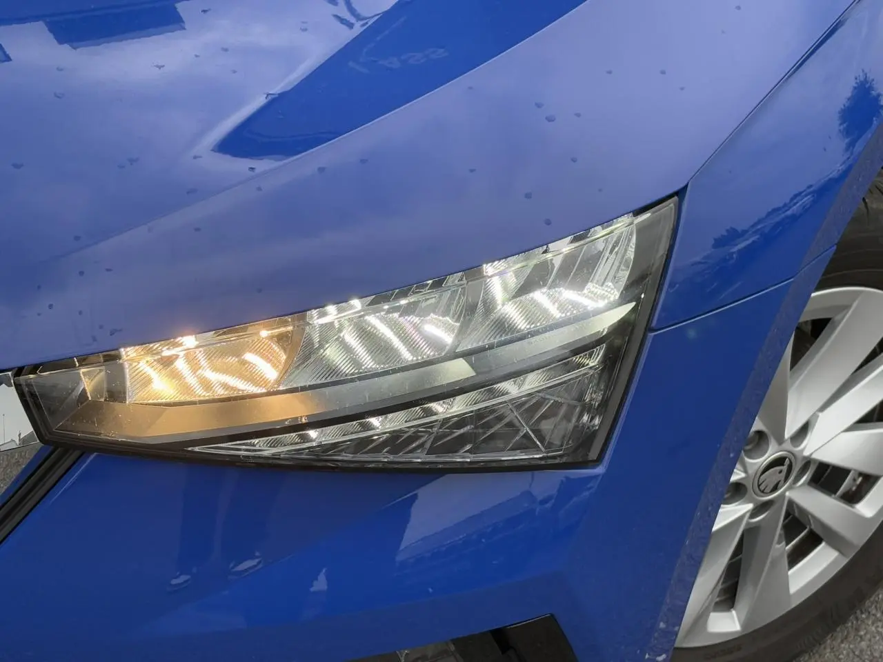 Gros plan sur le phare avant droit et la jante argentée d'une SKODA SCALA TSI 110 DSG Ambition bleu Energy 2024.