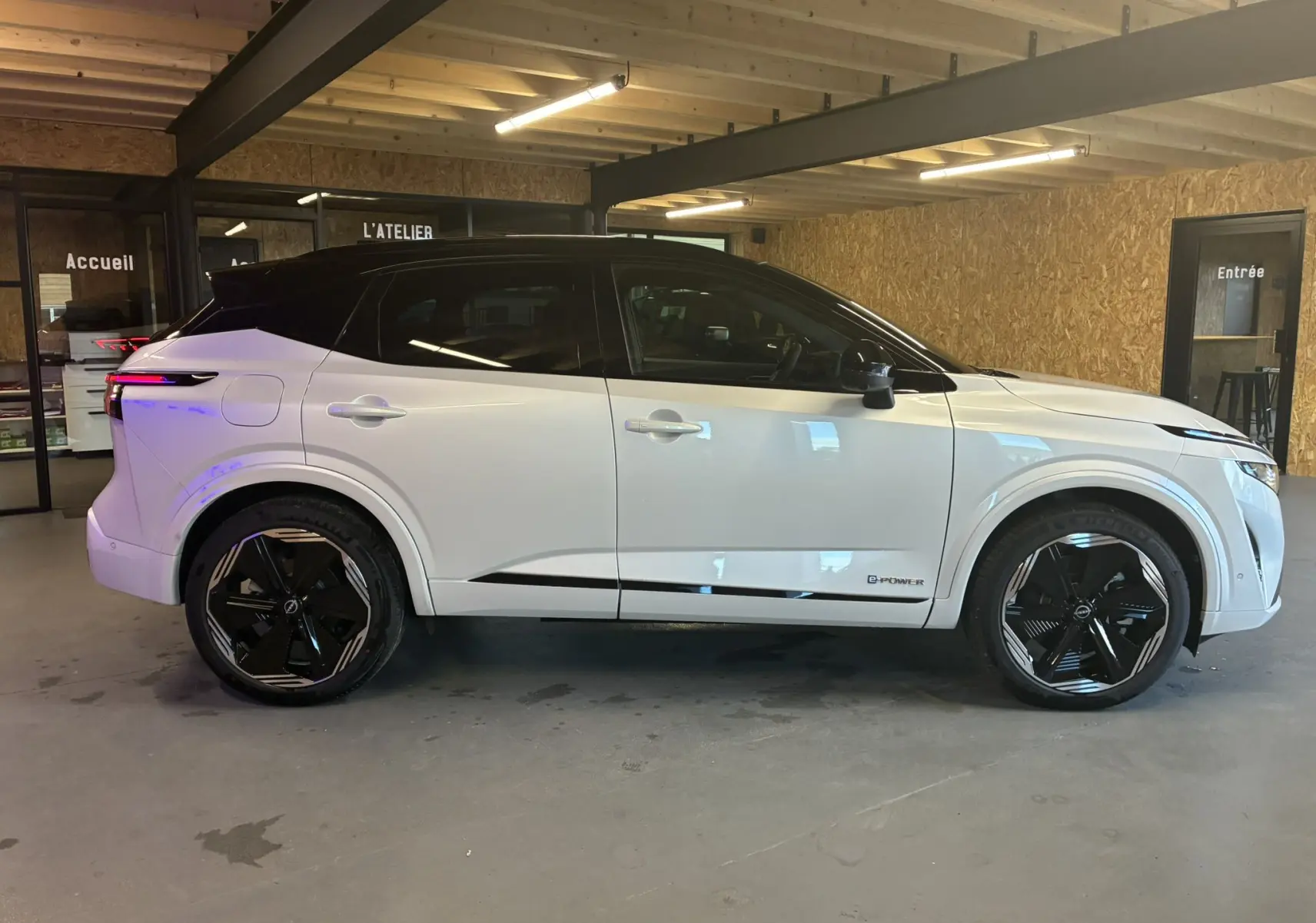 Profil côté droit du Nissan Qashqai 2025 blanc nacré avec toit noir et jantes noires en intérieur d'atelier.