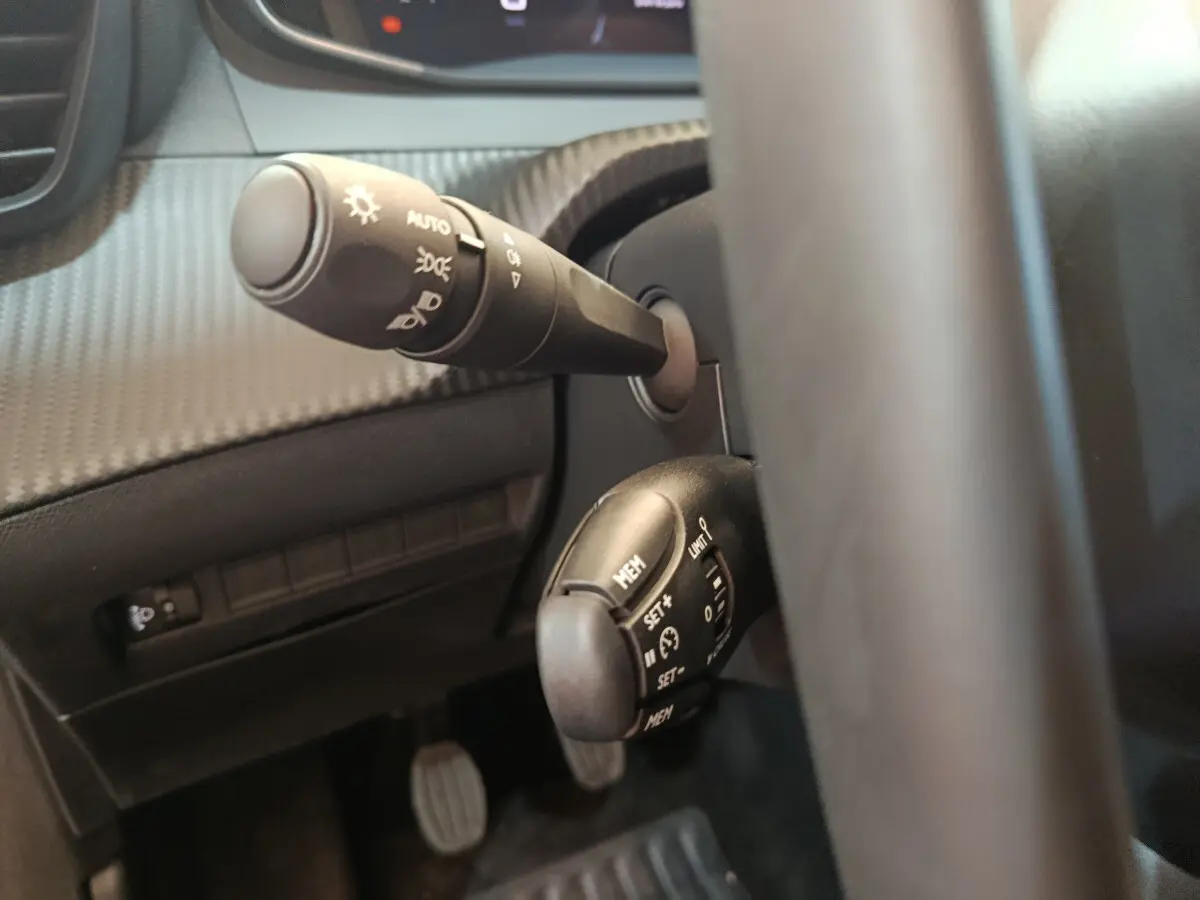 Gros plan sur les commandes au volant côté gauche d'une Peugeot 208 gris foncé, avec réglages phares et régulateur de vitesse.