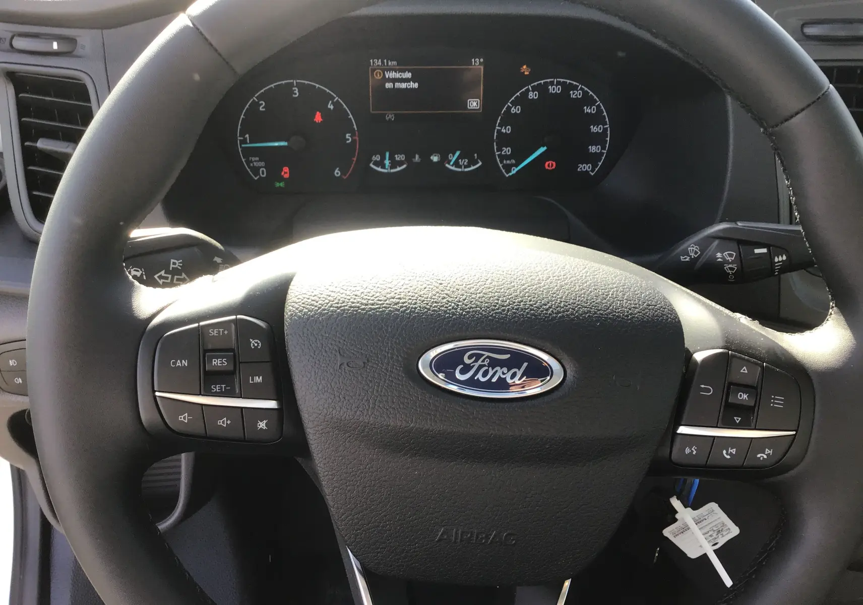 Vue rapprochée du volant noir Ford avec commandes intégrées et tableau de bord numérique en arrière-plan.