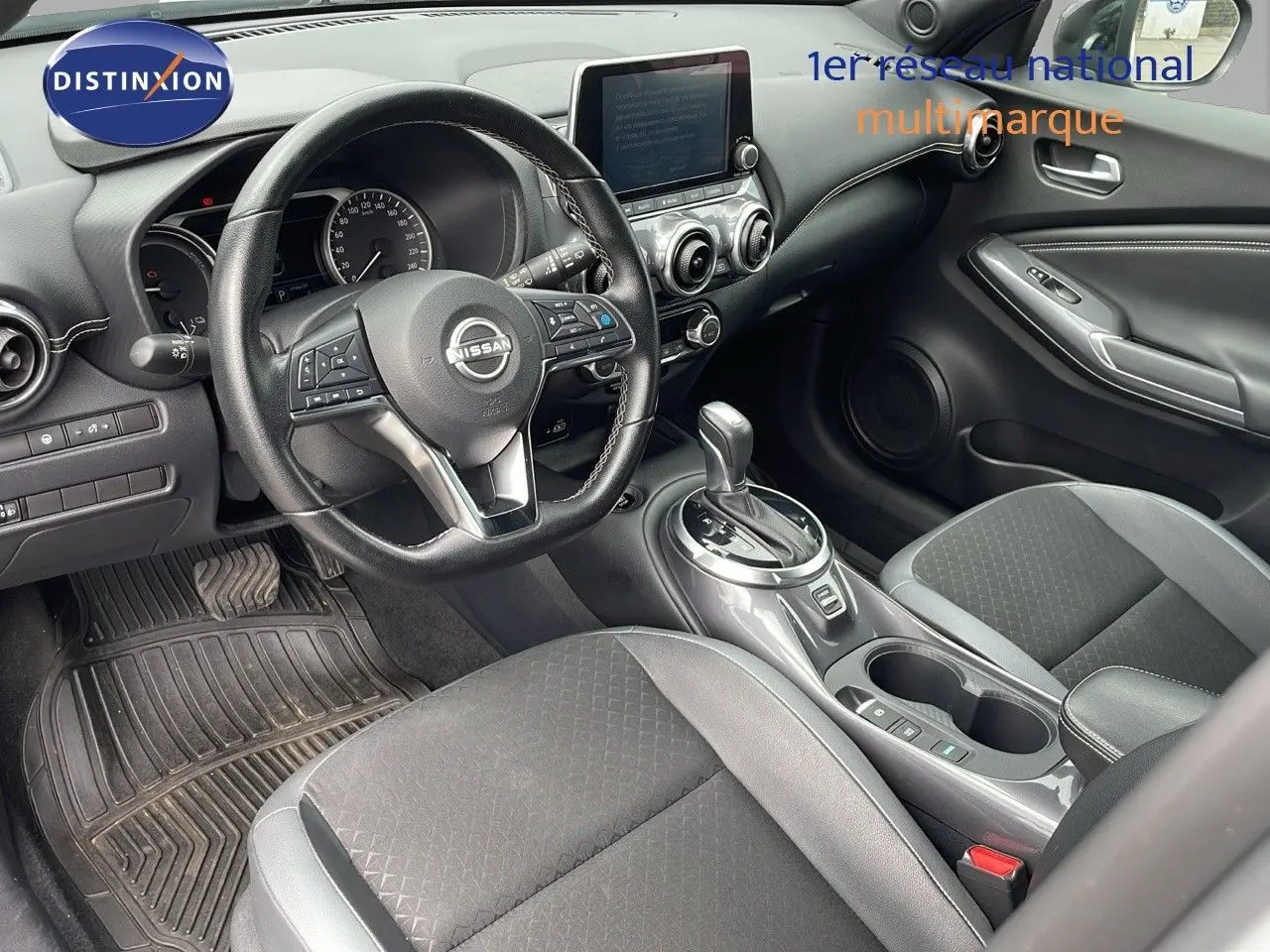 Intérieur du Nissan Juke Hybrid 2023 vu côté conducteur, avec volant cuir et console centrale grise brillante.
