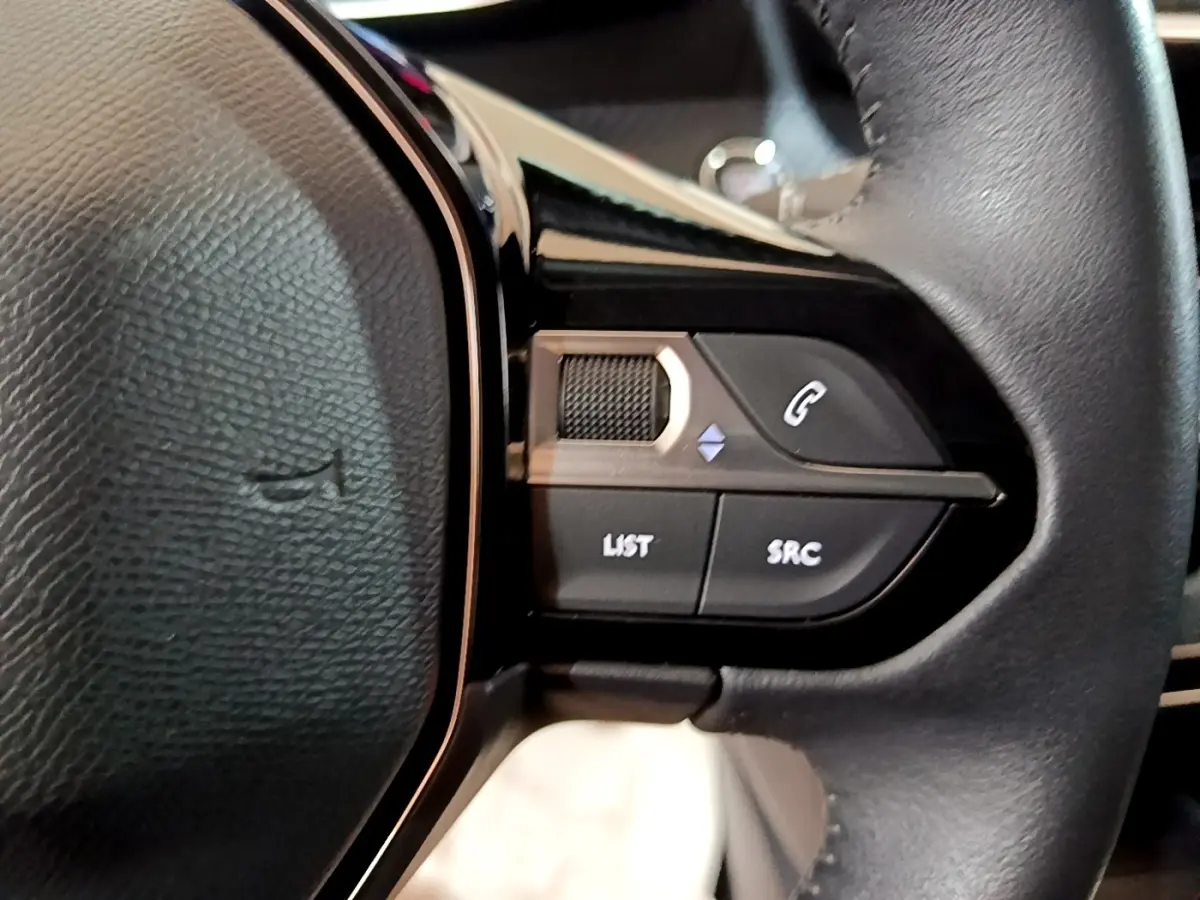 Gros plan sur les commandes multifonctions côté gauche du volant en cuir noir de la Peugeot 208 blanche.