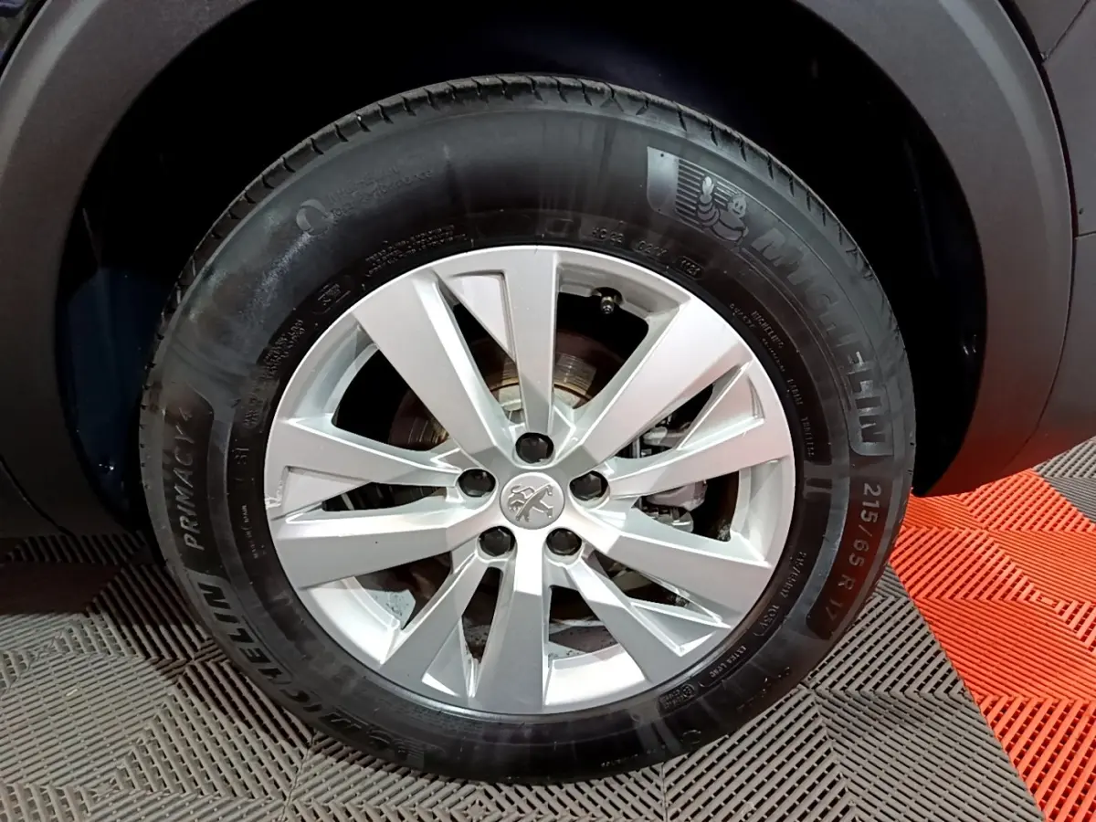 Gros plan sur la roue avant droite d’un Peugeot 5008 noir, jante alliage argentée avec logo lion au centre.