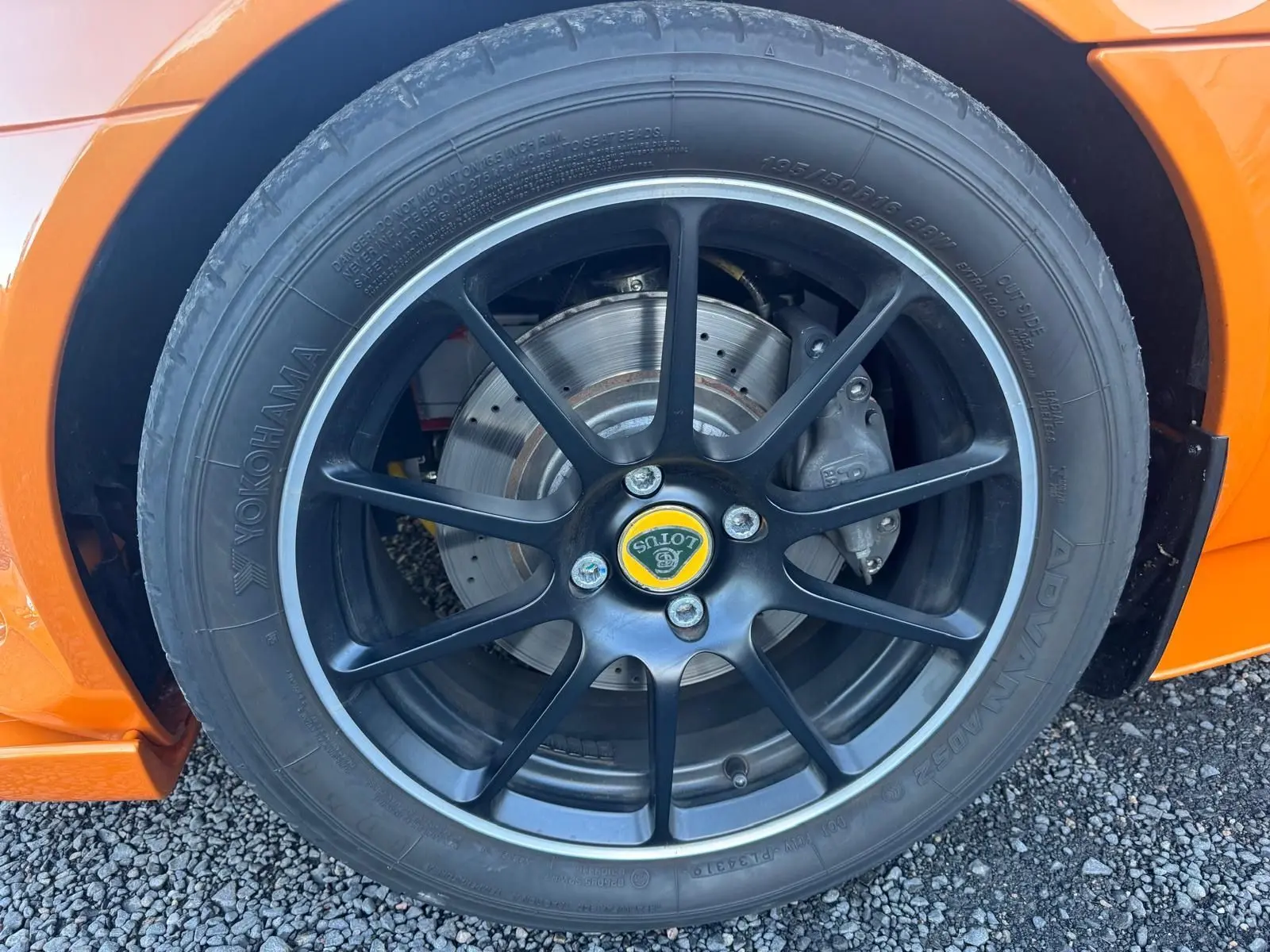 Gros plan sur la jante noire satinée forgée avec logo Lotus jaune, pneu Yokohama et partie orange de la carrosserie.