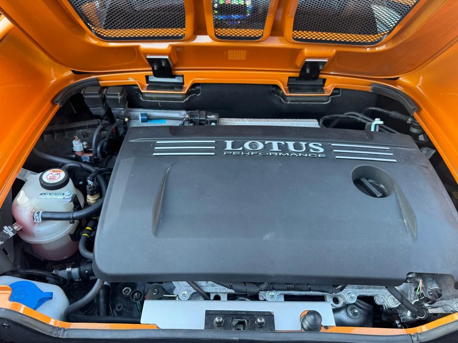 Vue rapprochée du compartiment moteur ouvert d'une Lotus Elise 3 Cup 250 orange, avec le cache moteur noir marqué "LOTUS PERFORMANCE".