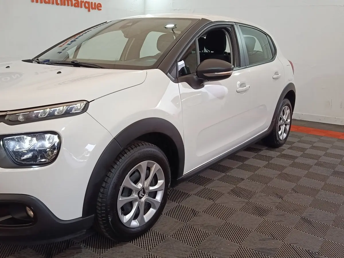 Vue 3/4 avant droit d'une Citroën C3 blanche avec phares distinctifs et jantes alliage en intérieur showroom.