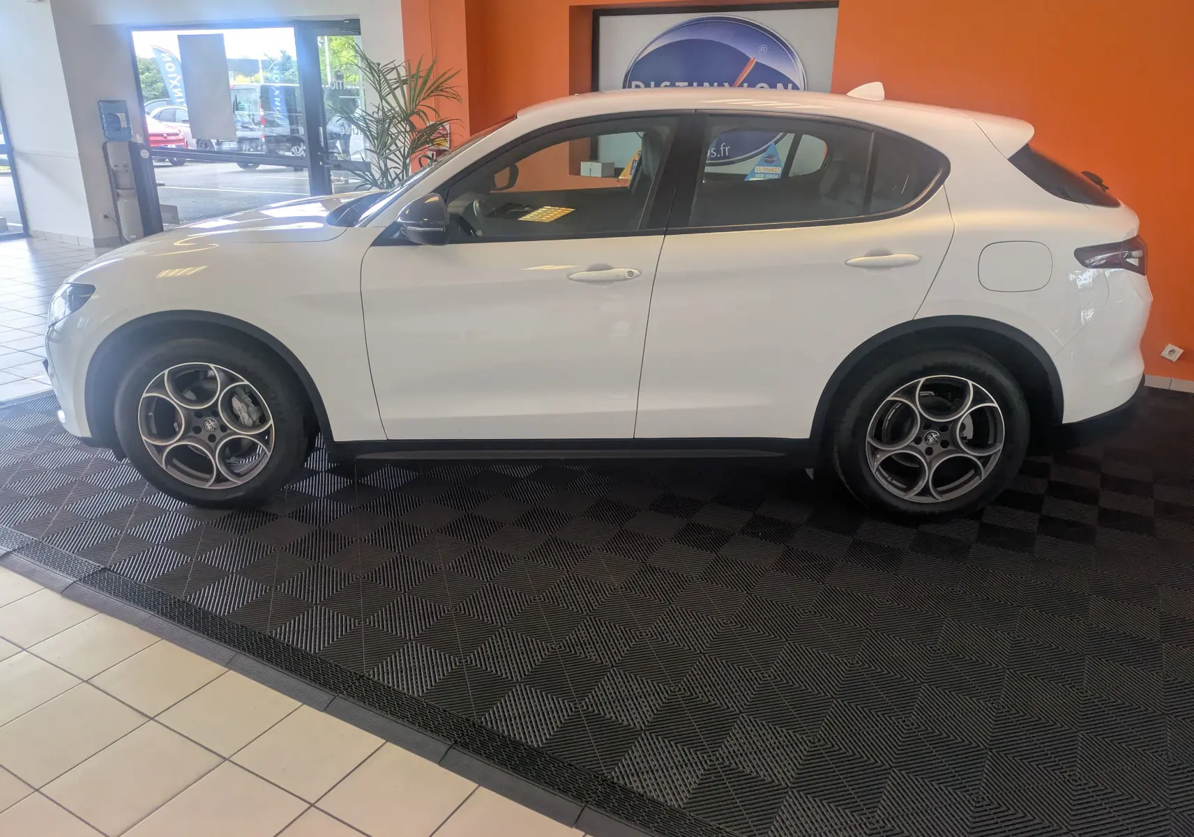 Profil côté gauche d'un Alfa Romeo Stelvio blanc 2023 exposé en intérieur avec un mur orange en arrière-plan.