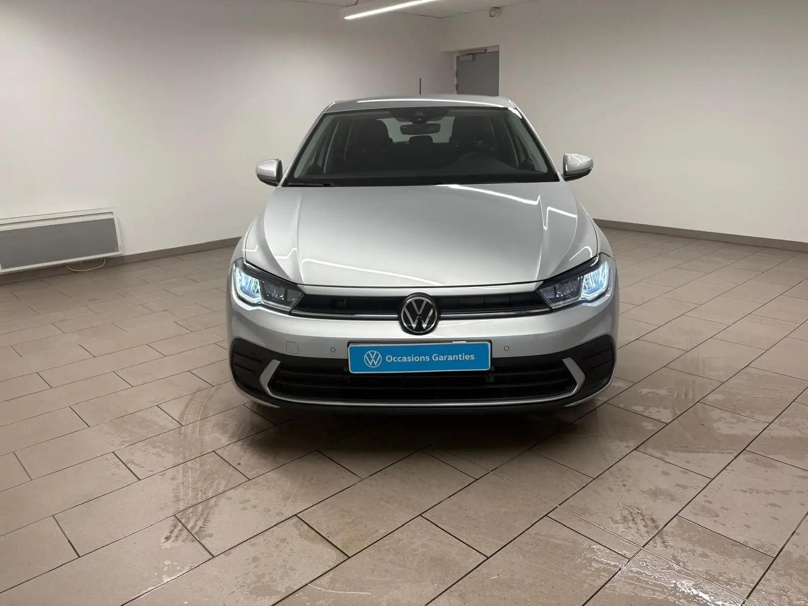 Vue frontale d'une Volkswagen Polo 2024 en peinture métallisée reflet d'argent avec phares allumés dans un showroom.