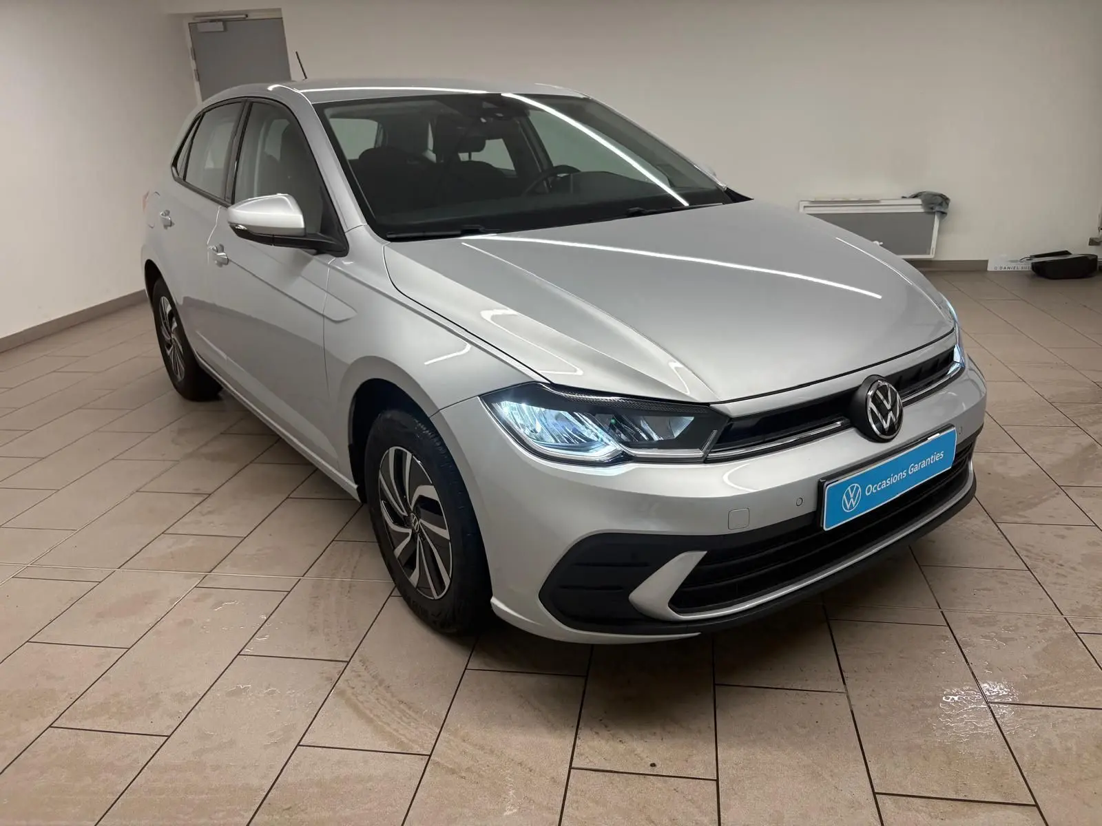 Volkswagen Polo 2024 Reflet d'argent vue 3/4 avant droit avec phares allumés en intérieur showroom.