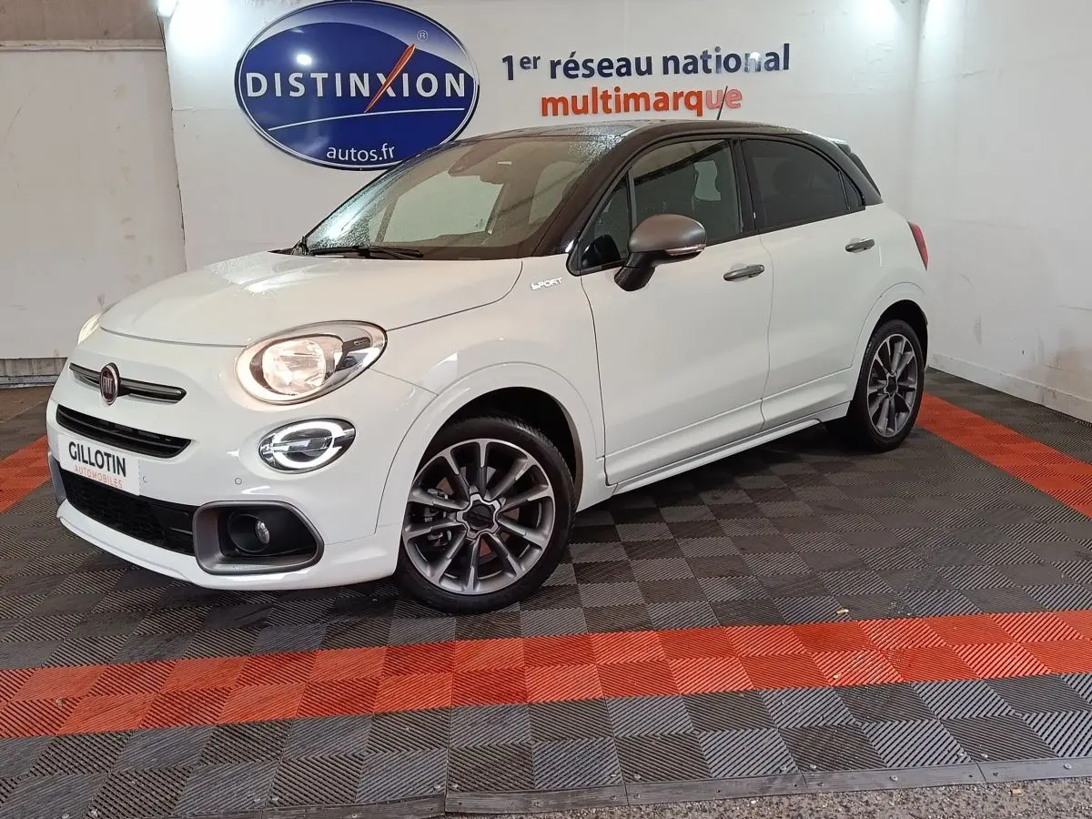 Fiat 500X blanc à toit noir en 3/4 avant droit, avec jantes sport et feux allumés dans un showroom.