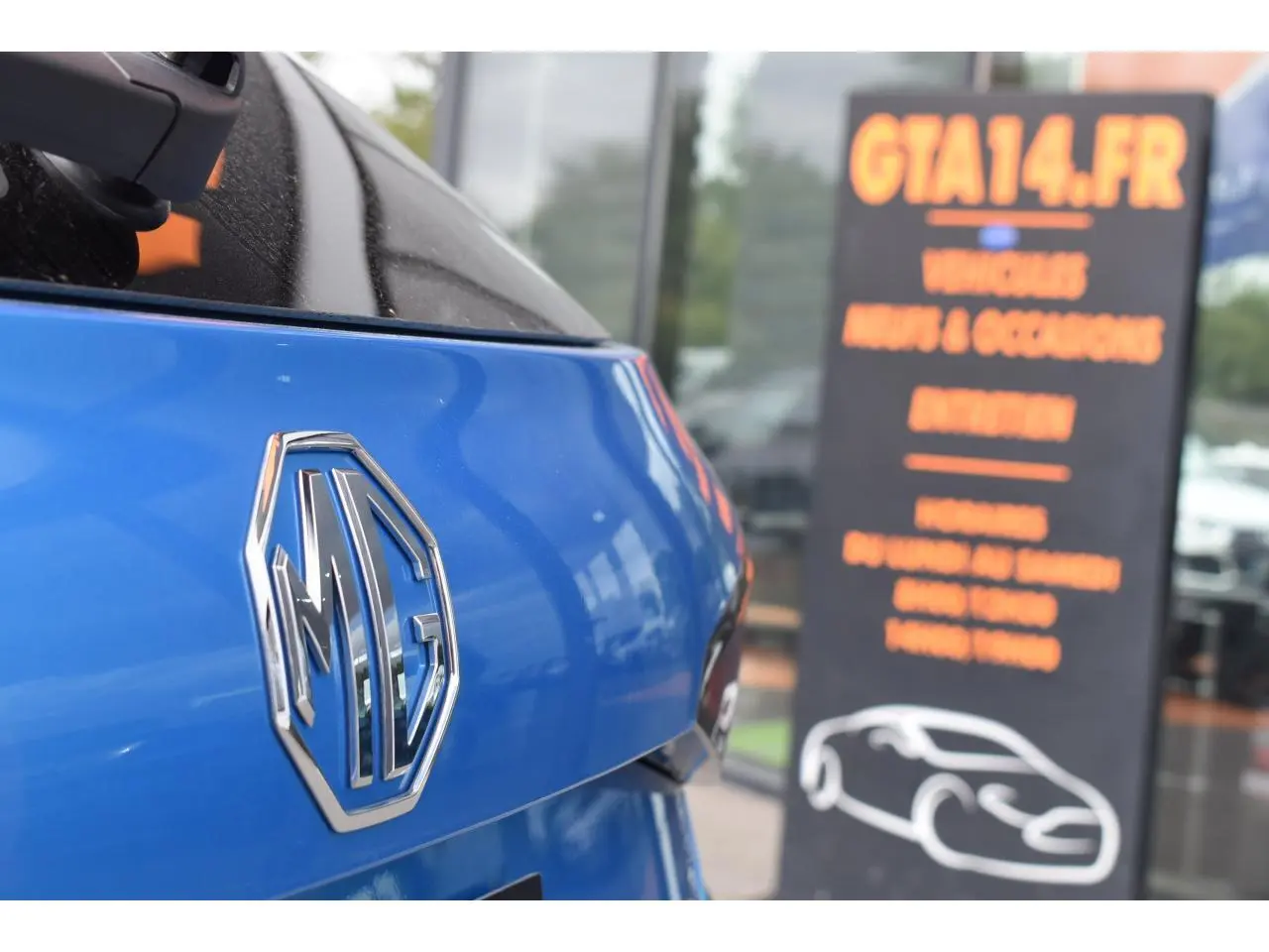 Gros plan sur le logo MG à l'arrière d'une MG3 1.5 HYBRID+ 195 Luxury bleu métallisé, avec reflet du ciel et panneau flou en arrière-plan.