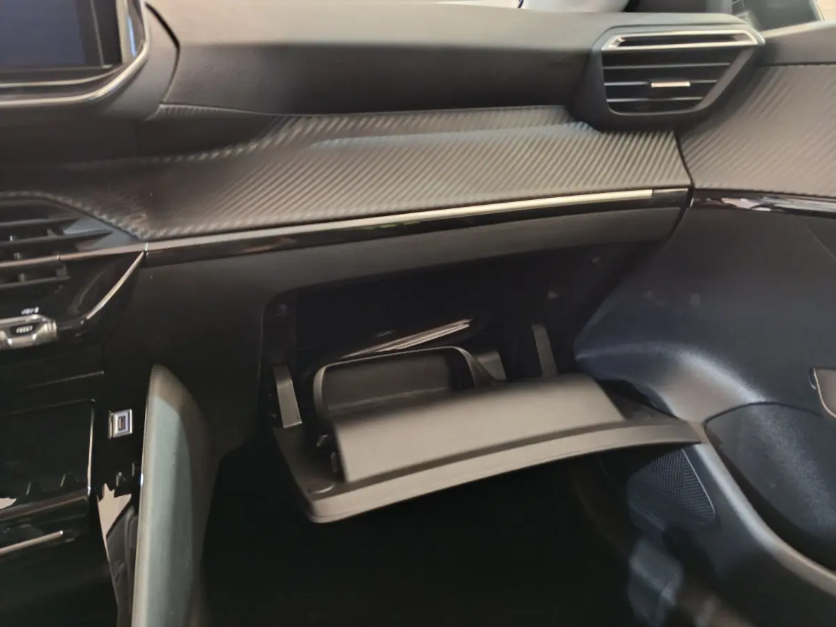 Vue intérieure du tableau de bord gris foncé de la Peugeot 208 2025 avec boîte à gants ouverte et rangement visible.