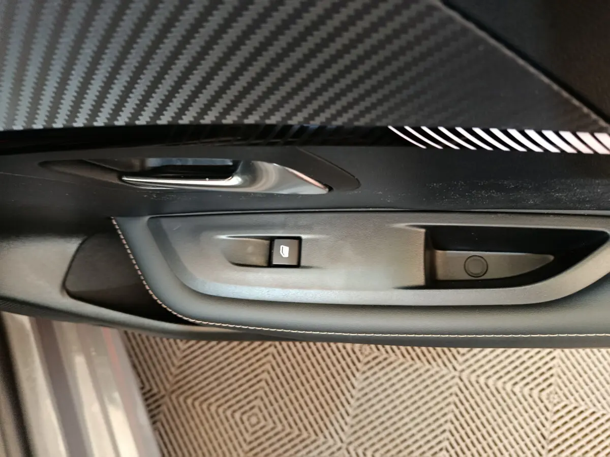 Gros plan sur la commande de lève-vitre côté conducteur de la Peugeot 208 gris foncé, avec insert décoratif en fibre de carbone.