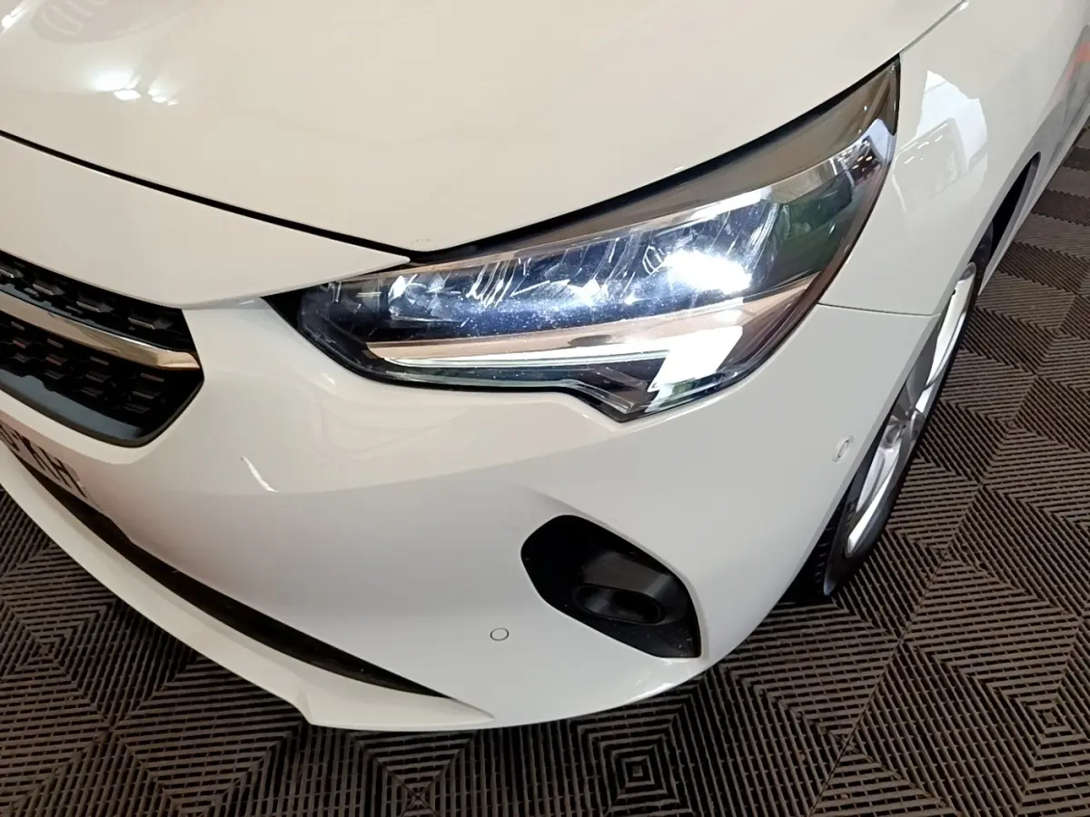 Vue 3/4 avant droit d'une Opel Corsa blanche 2023, mettant en valeur le phare LED et le pare-chocs avec capteurs de stationnement.