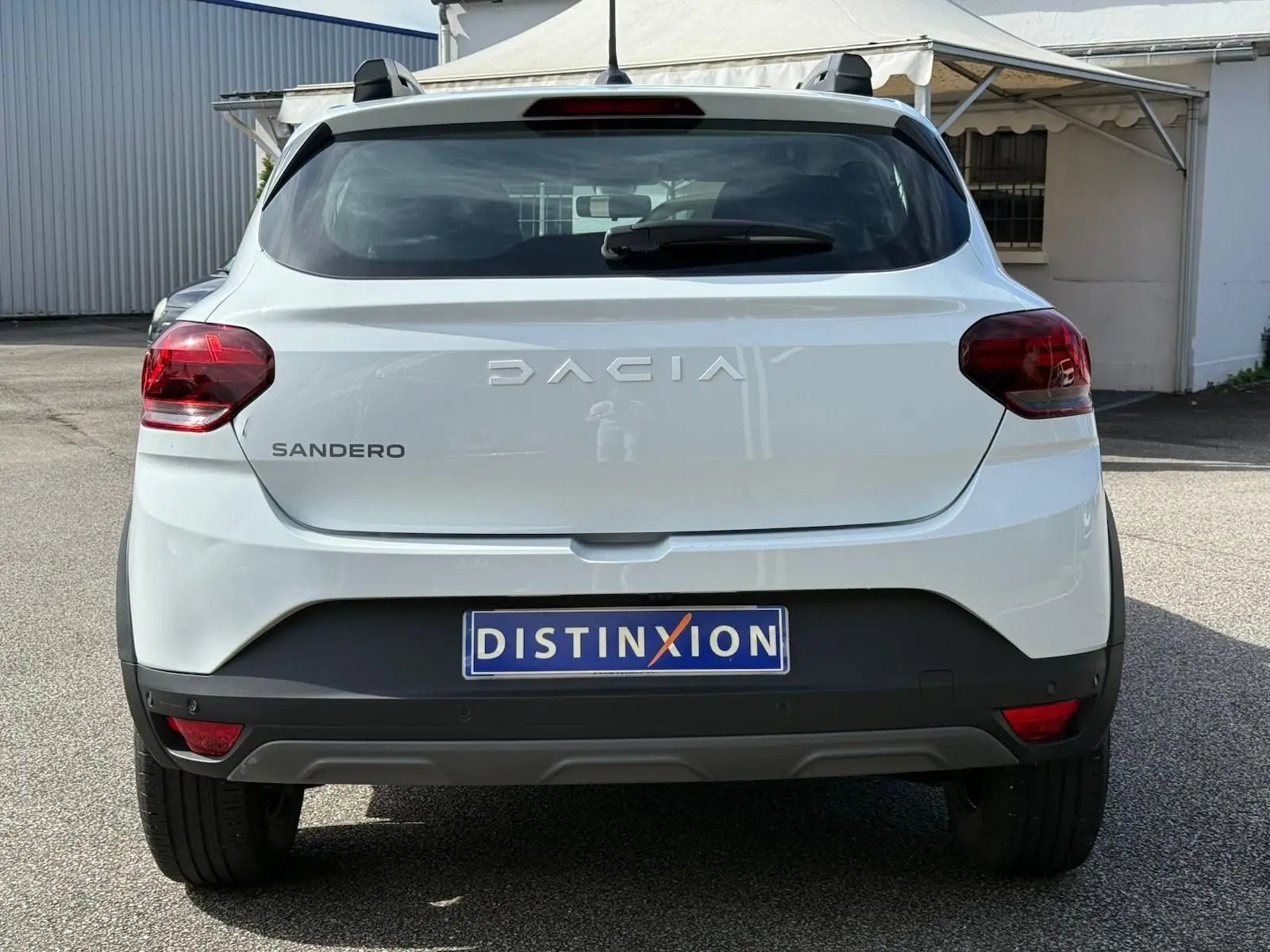 Vue arrière d'une Dacia Sandero blanche 2023 avec logo distinctif et feux arrière rouges sur parking extérieur.