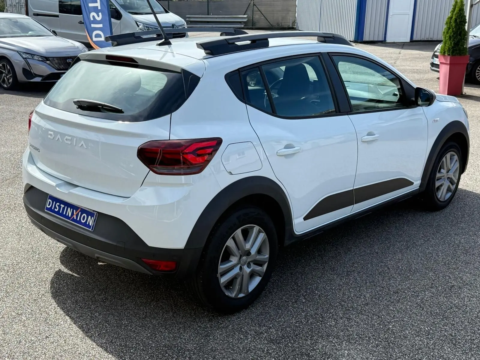 Vue 3/4 arrière droite d'une Dacia Sandero blanche 2023 avec protections noires et toit ouvrant visible.