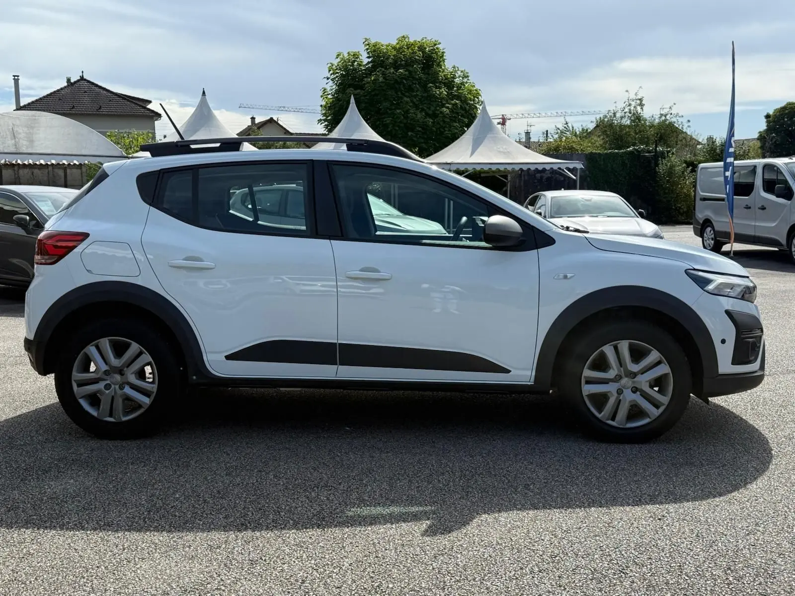 Profil côté gauche d'une Dacia Sandero blanche 2023 avec protections noires et barres de toit sur parking extérieur.