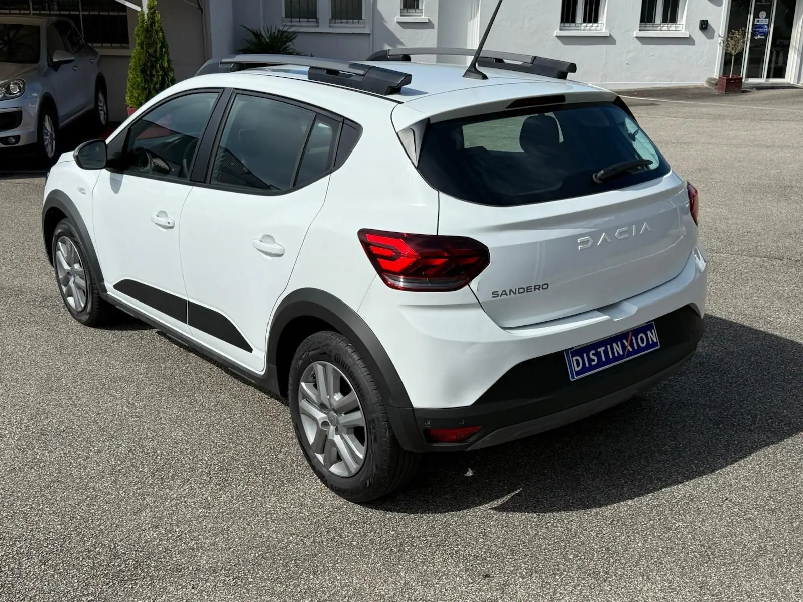 Vue 3/4 arrière droite d'une Dacia Sandero blanche 2023 avec toit ouvrant et protections noires sur les passages de roues.