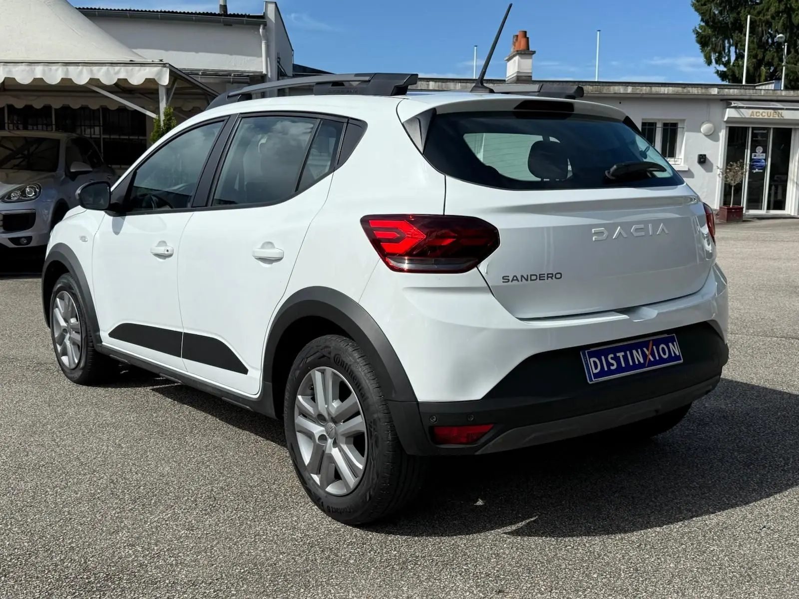 Vue 3/4 arrière droite d'une Dacia Sandero blanche 2023 avec barres de toit et détails noirs contrastants.