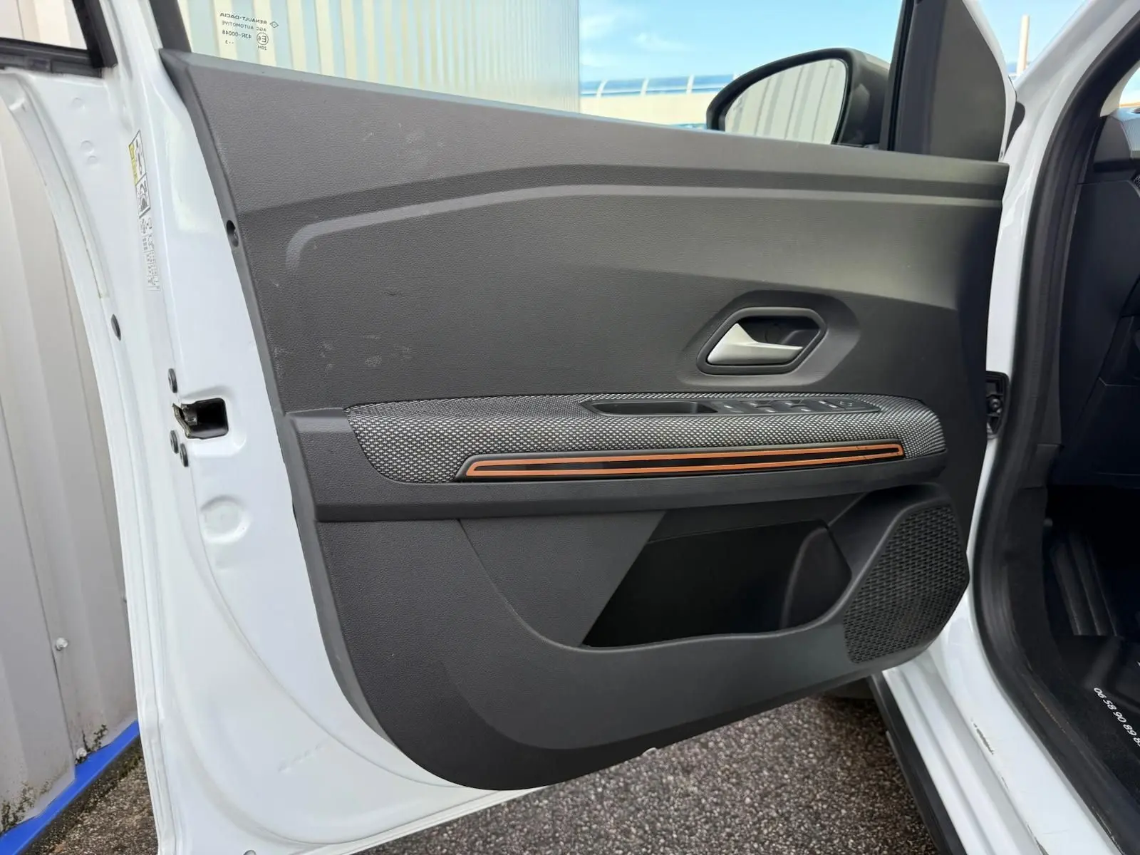 Intérieur de la porte avant gauche blanche d'une Dacia Sandero 2023 avec bande décorative orange et haut-parleur intégré.