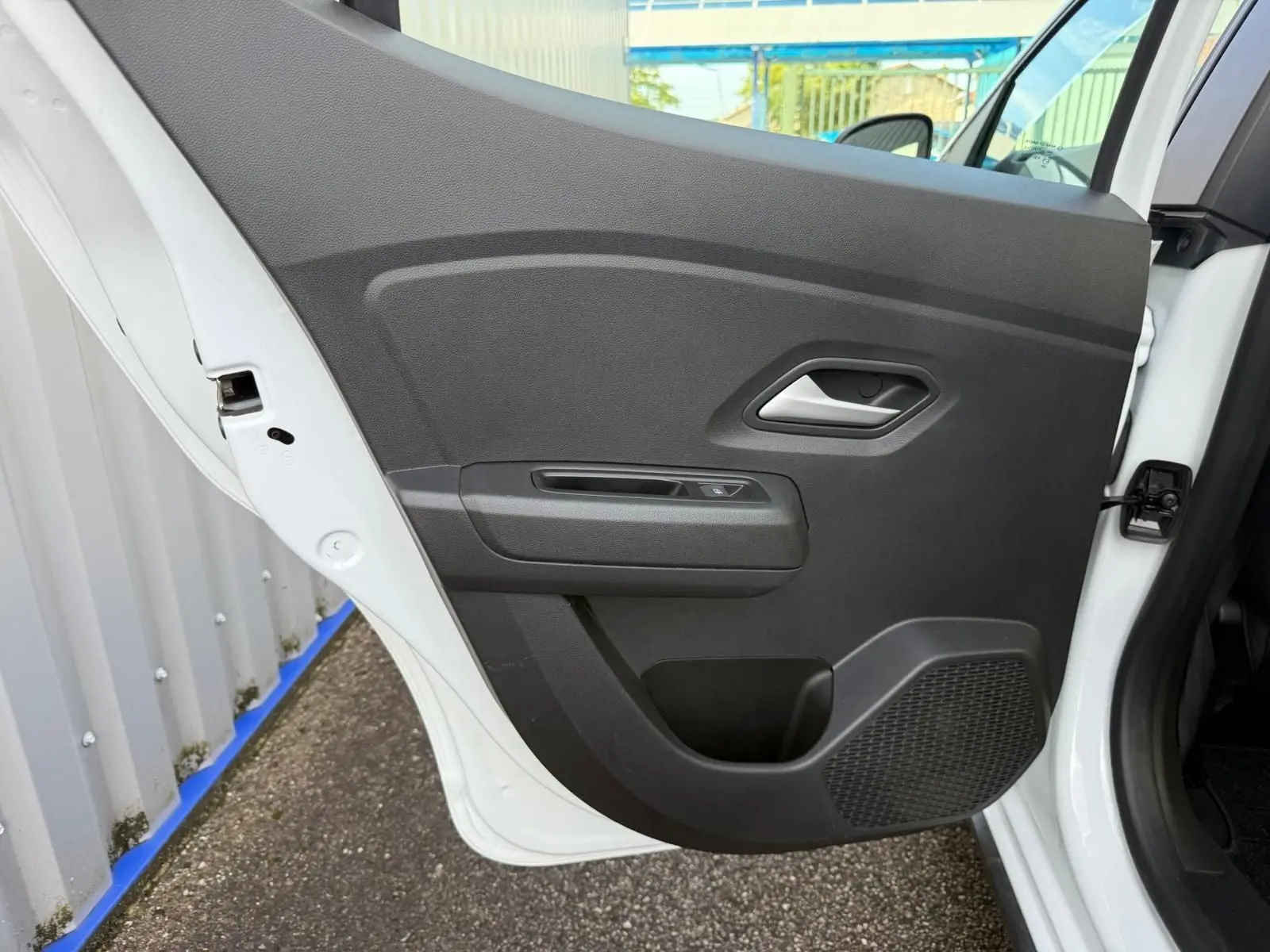 Porte arrière gauche ouverte d'une Dacia Sandero blanche 2023, montrant le panneau intérieur noir avec poignée et haut-parleur intégré.