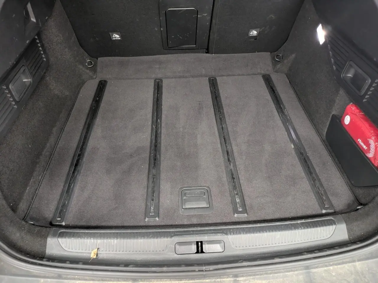 Coffre vu de dessus du Citroën C5 X 2024, tapis gris foncé avec rails noirs et trousse de secours rouge sur le côté droit.