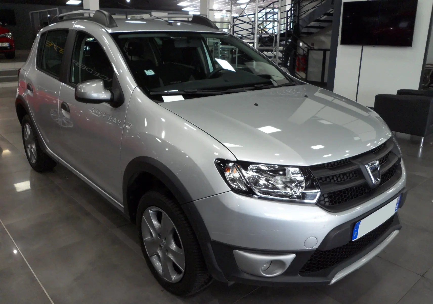 Vue 3/4 avant d'une Dacia Sandero Stepway gris platine en showroom, avec jantes alliage et barres de toit noires.