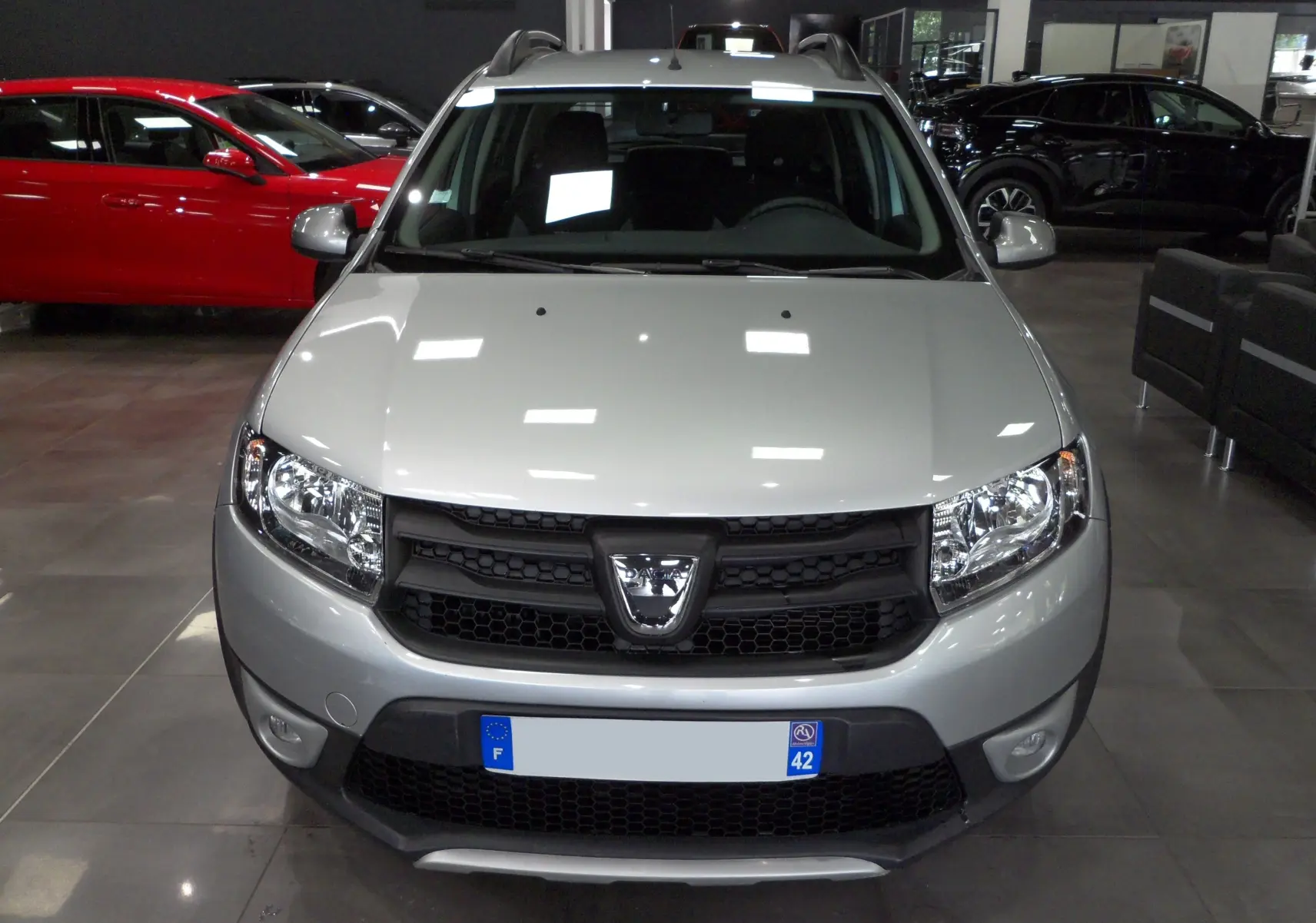 Vue frontale d'une Dacia Sandero Stepway TCe 90 gris platine, avec calandre noire et phares allumés en showroom.
