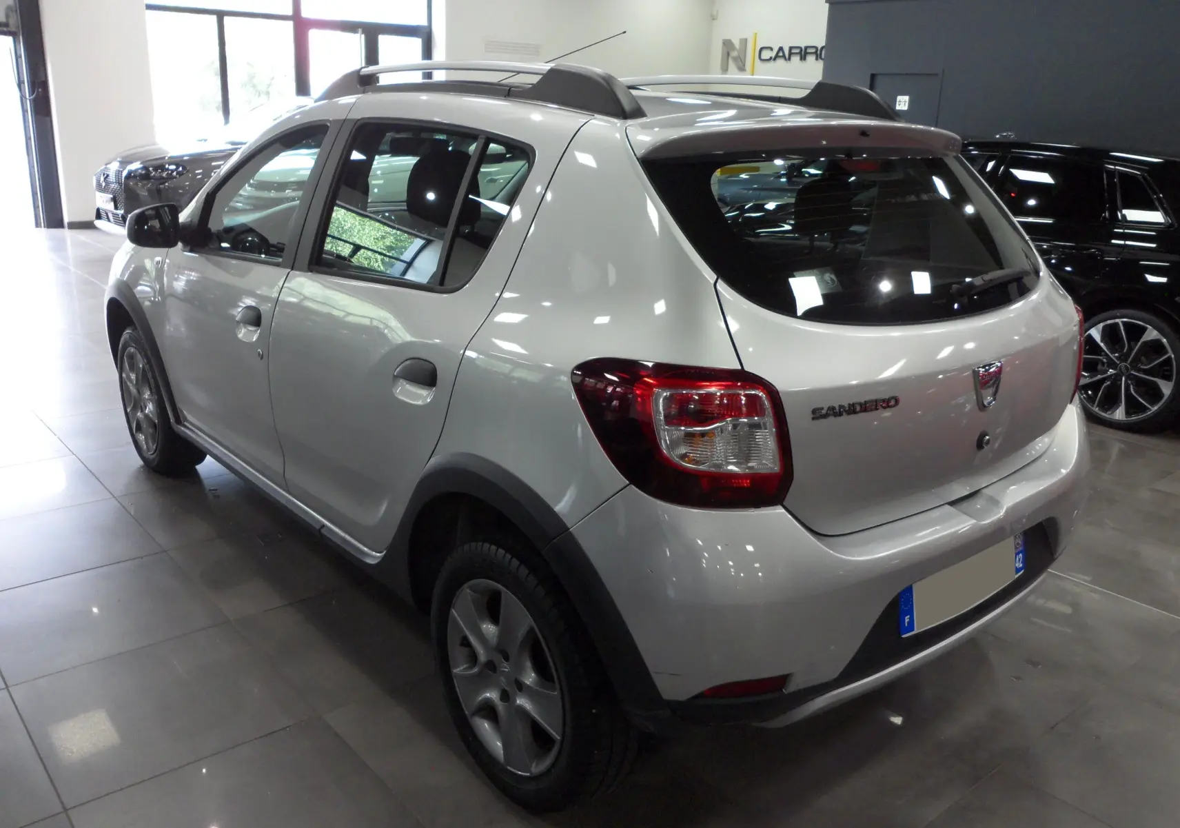Vue 3/4 arrière droite d'une Dacia Sandero Stepway gris platine en intérieur, avec barres de toit et jantes alliage.