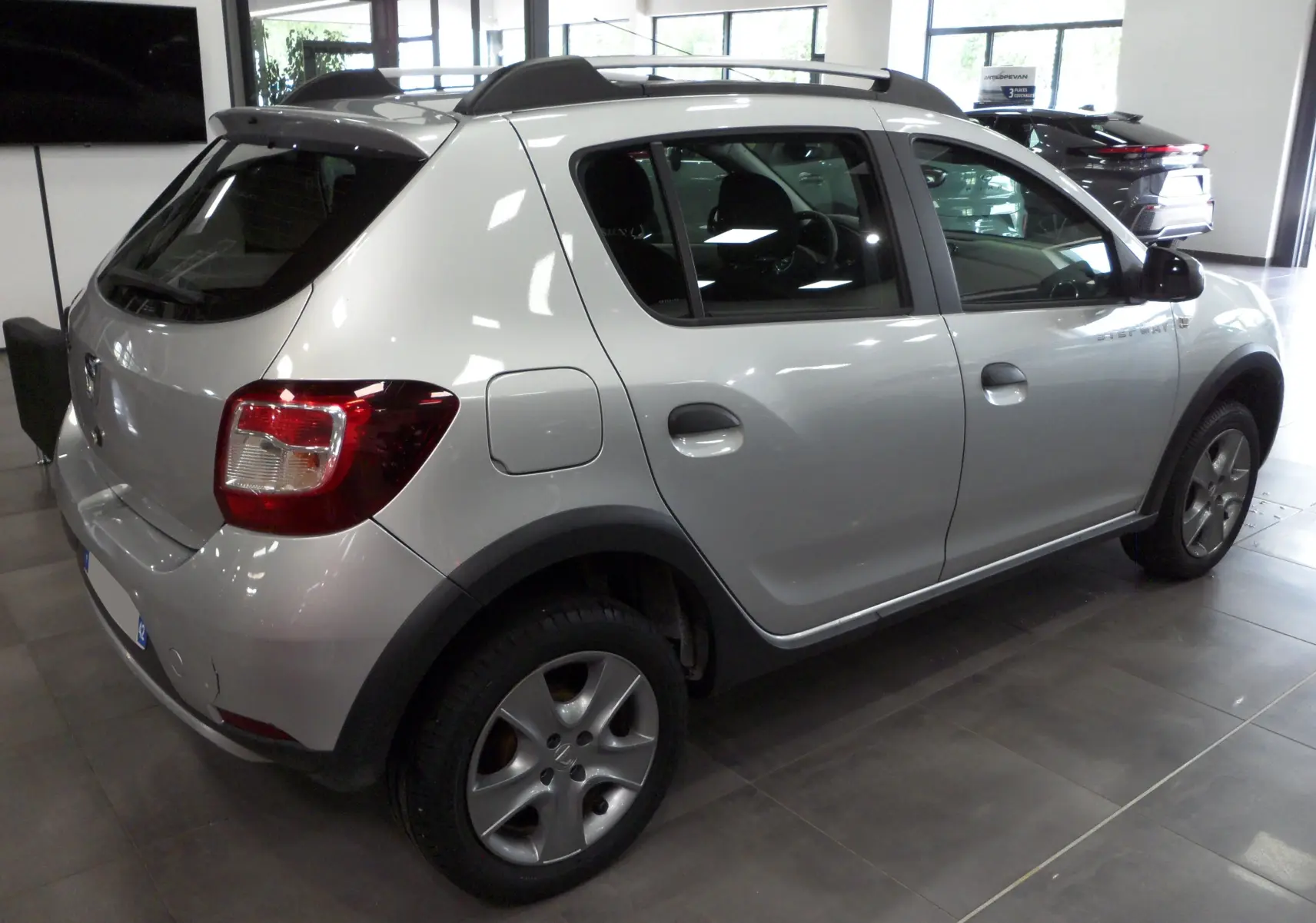 Vue 3/4 arrière droite d'une Dacia Sandero Stepway gris platine en intérieur, avec jantes alliage et barres de toit noires.