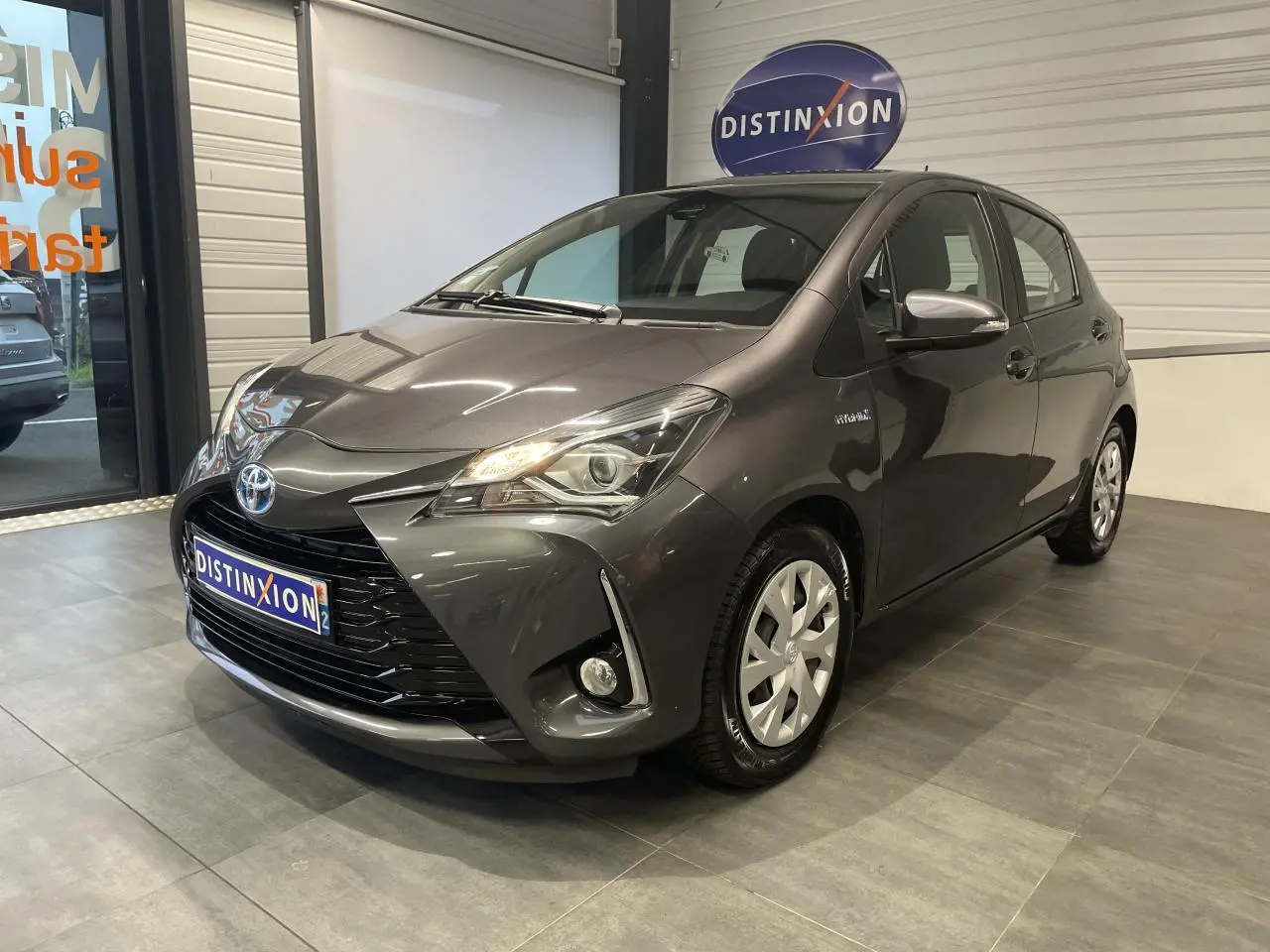 Toyota Yaris hybride gris foncé vue 3/4 avant droit en intérieur avec calandre noir laqué et phares allumés.