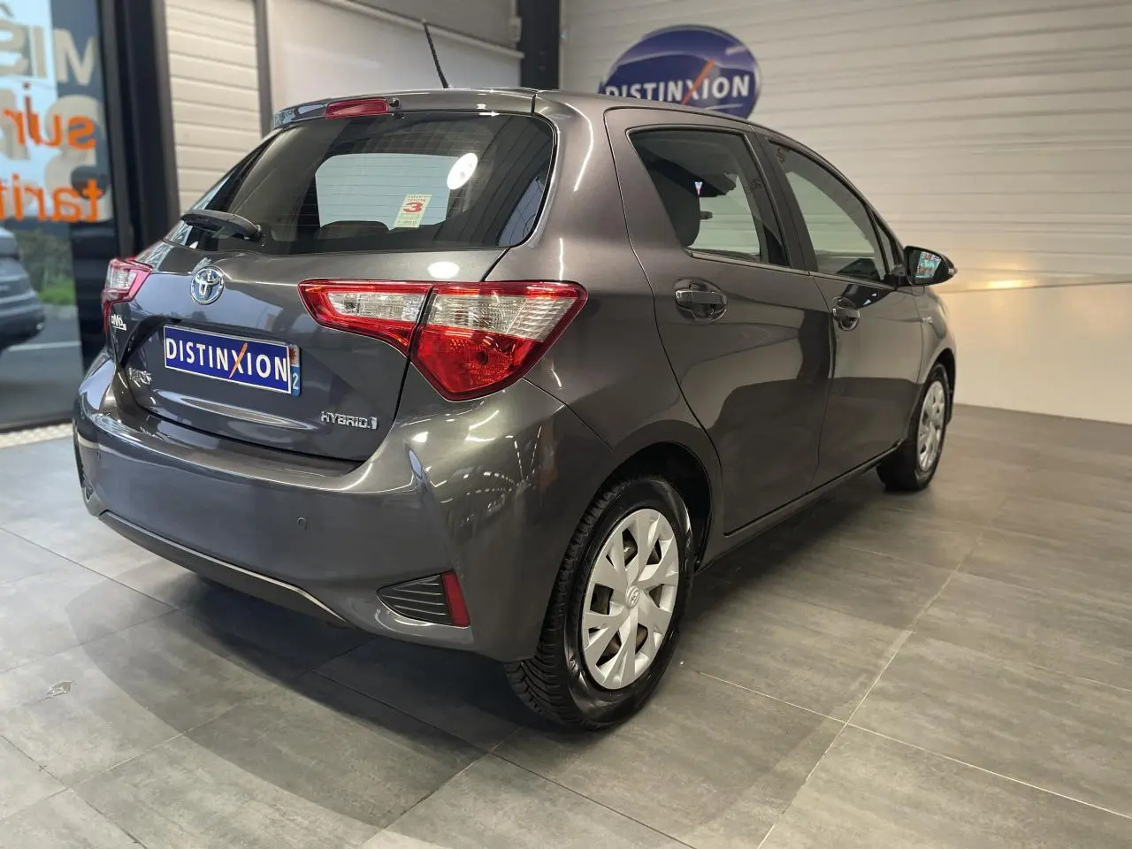 Toyota Yaris hybride gris foncé vue 3/4 arrière droit en intérieur avec jantes à enjoliveurs blancs.