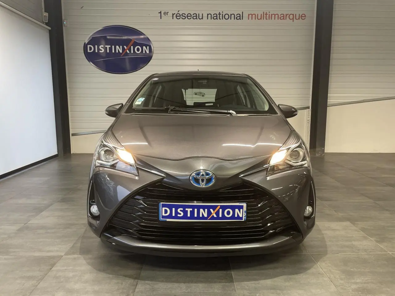 Vue frontale d'une Toyota Yaris hybride gris foncé métallisé avec calandre noir laqué et phares allumés.