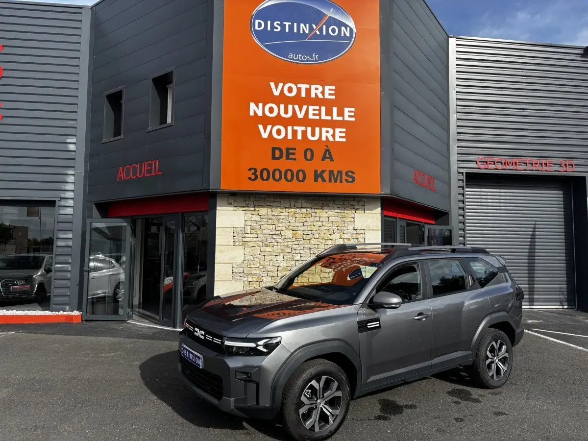SUV Dacia Bigster gris foncé vu en 3/4 avant droit devant un bâtiment commercial avec enseigne orange.