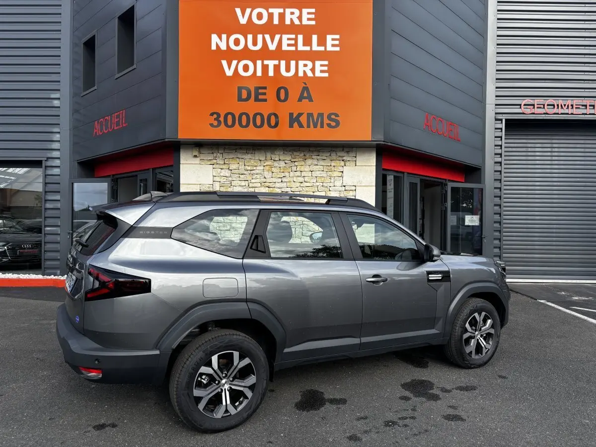 Vue de profil côté gauche d'un Dacia Bigster gris foncé avec rails de toit noirs garé devant un bâtiment commercial.