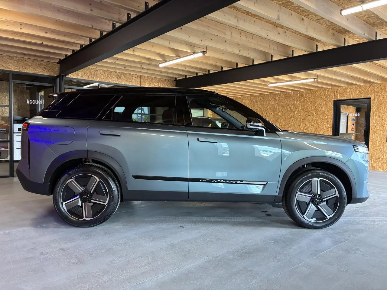 SUV JAECOO J7 gris olive avec toit noir, vu de profil côté gauche dans un garage en bois.